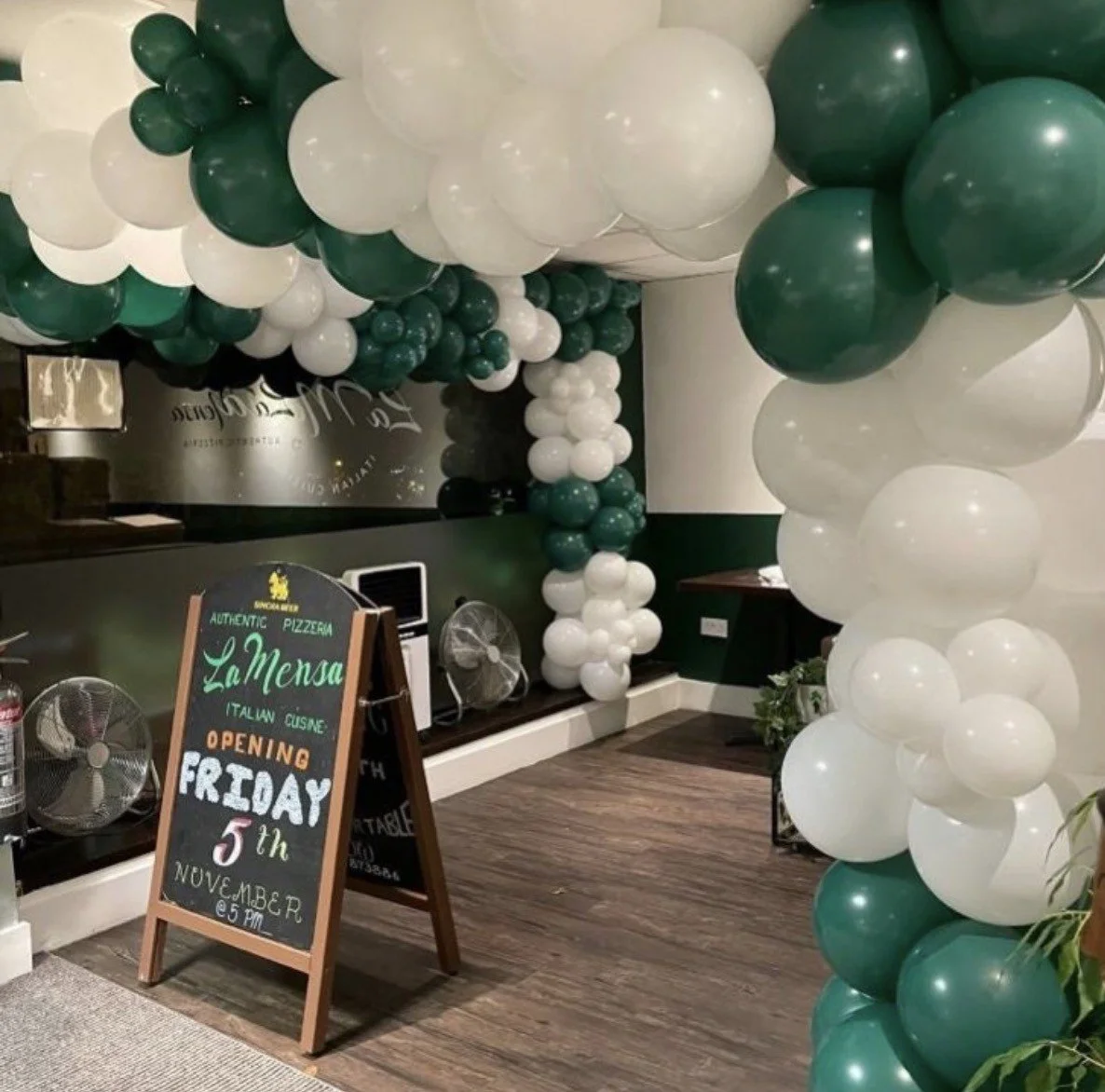 A restaurant entrance decorated with green and white balloon arches, with a chalkboard sign announcing the opening of La Mensa Italian Cuisine on Friday, November 5th at 5 PM.