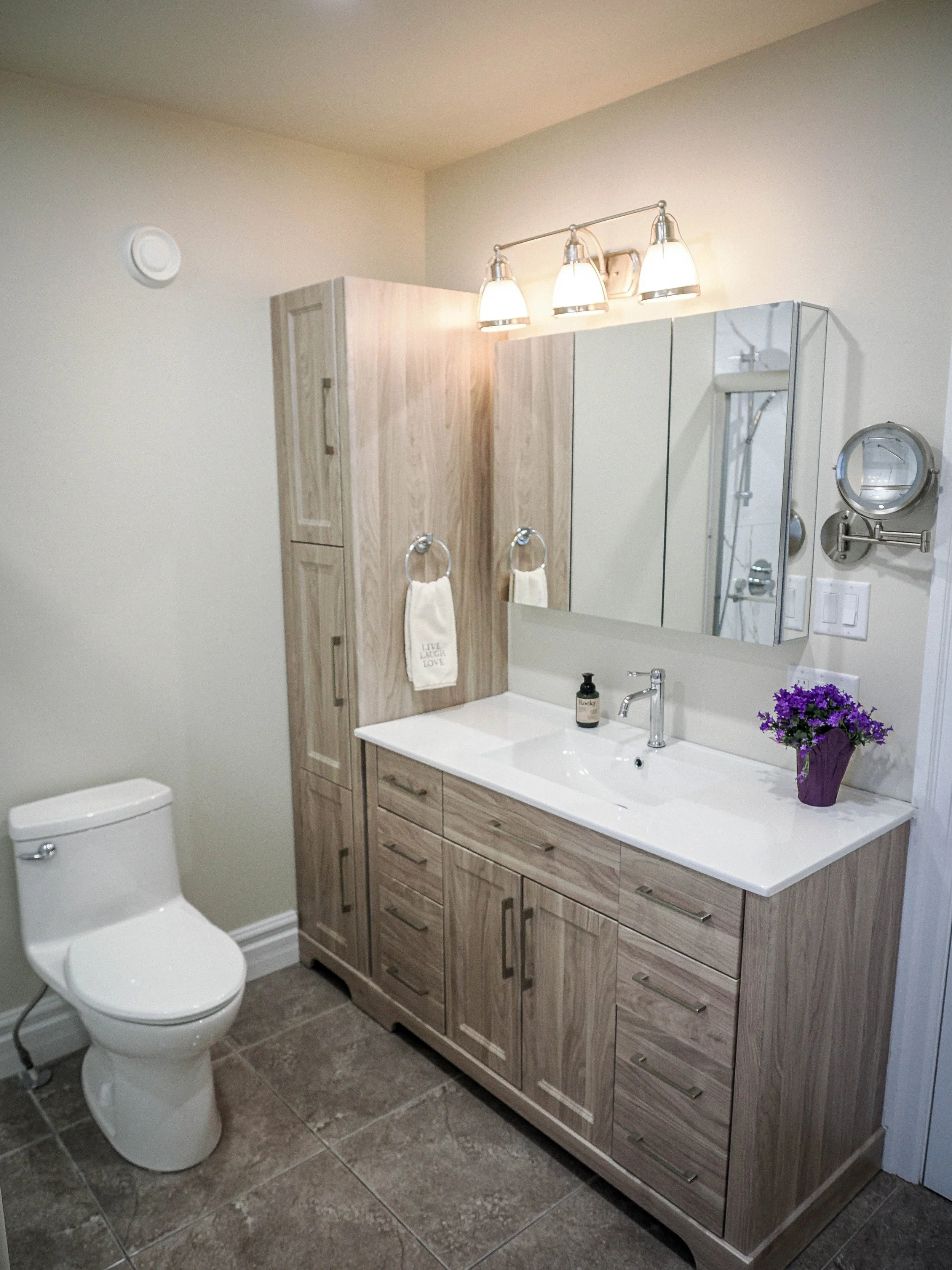 A double-drawer vanity with a quartz countertop, paired with a tall matching linen tower for extra storage. A tri-fold mirrored medicine cabinet and modern three-light vanity fixture complete the space, along with a swing-arm magnifying mirror for ad