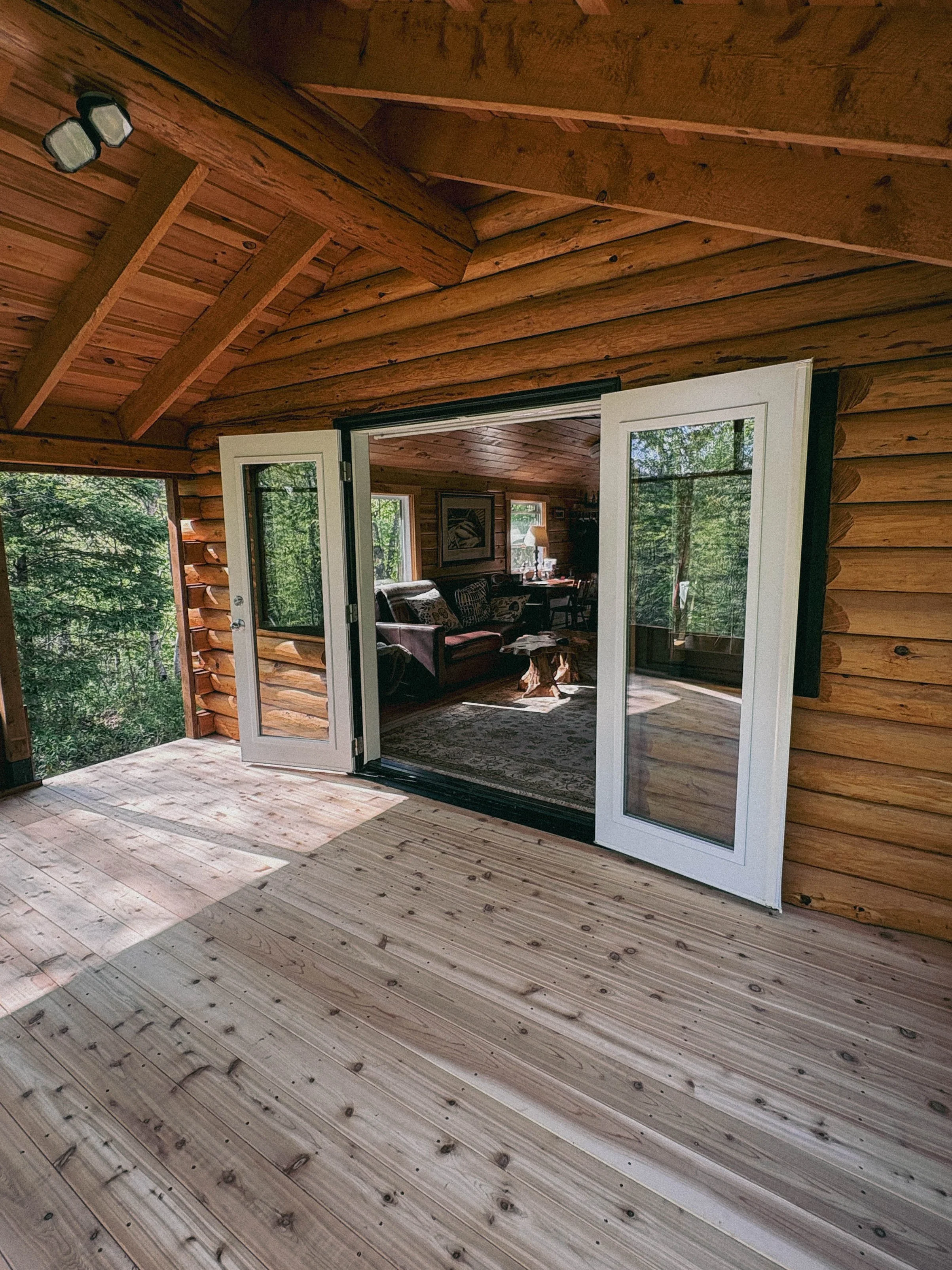 A screened, timber-framed porch addition with tongue-and-groove wood ceiling and a glass-panel door connecting seamlessly to the main cottage, surrounded by forest views.