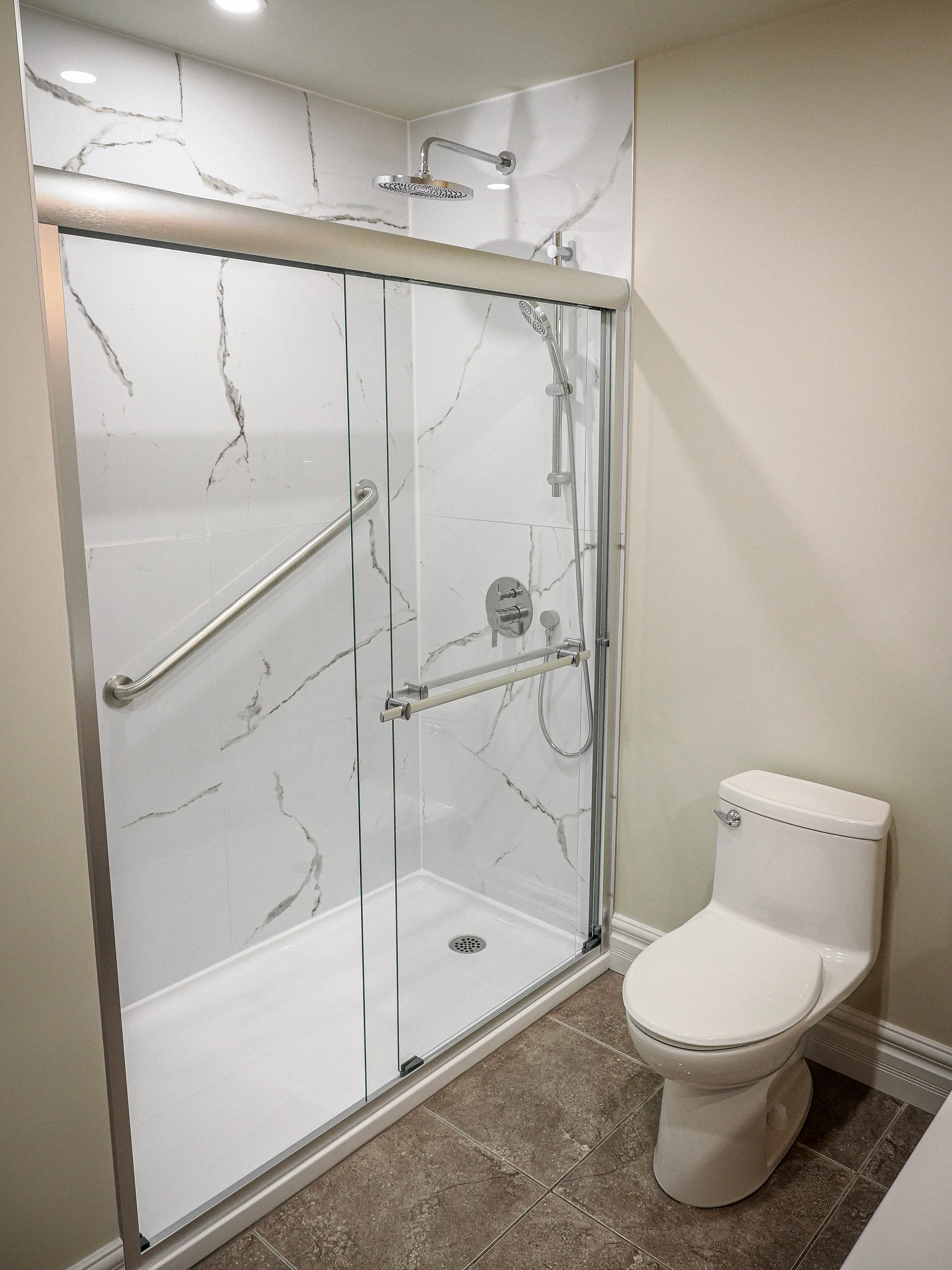 A complete bathroom remodel featuring a walk-in shower with sleek marble-look porcelain tile, a rainfall showerhead with handheld wand, and a frameless sliding glass door. Thoughtful details like the built-in safety grab bar and bench-height seat add