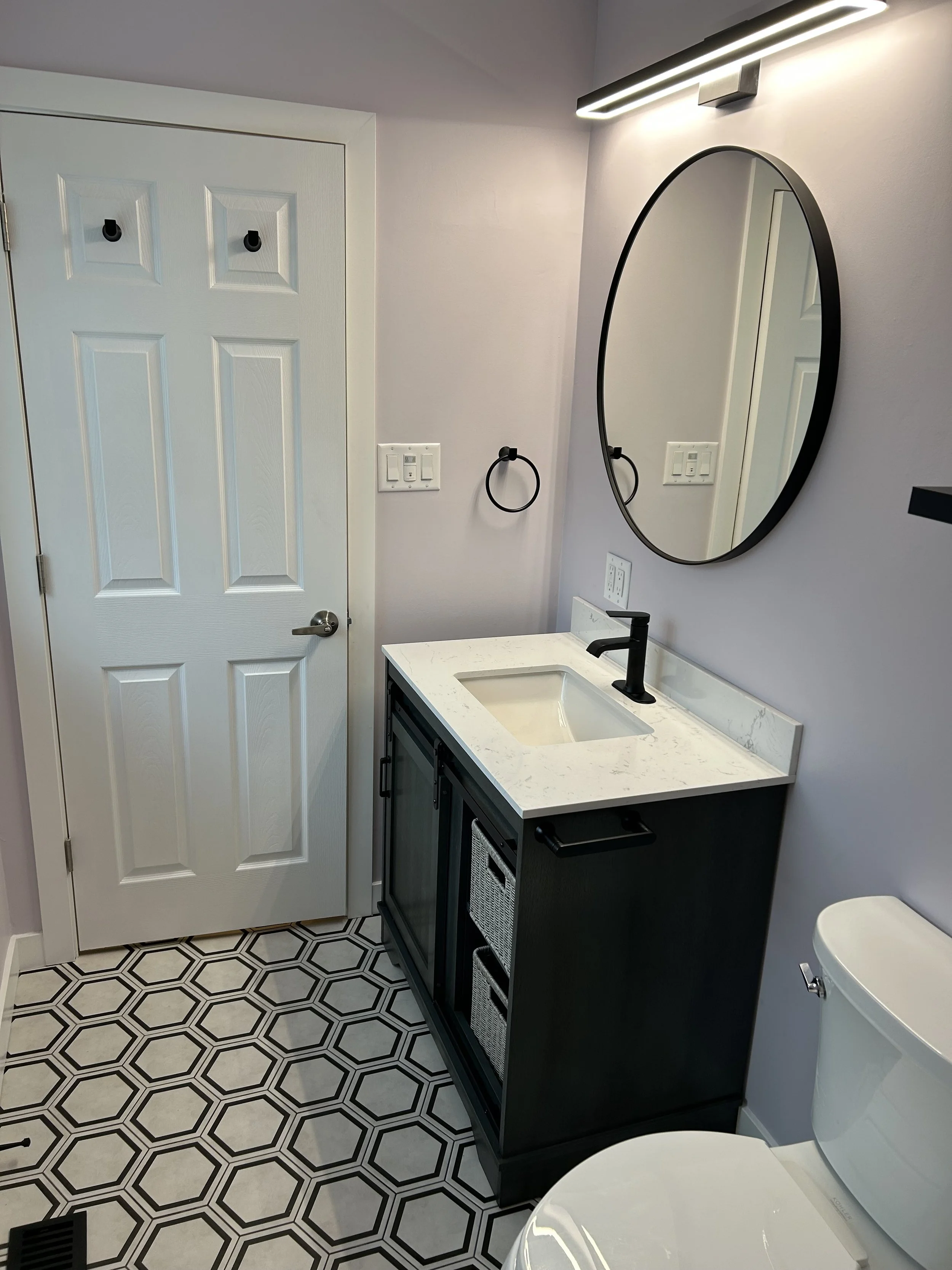 A modern bathroom with a black vanity, white countertop, round mirror, black faucet, hexagon patterned floor, white walls, and a white door.