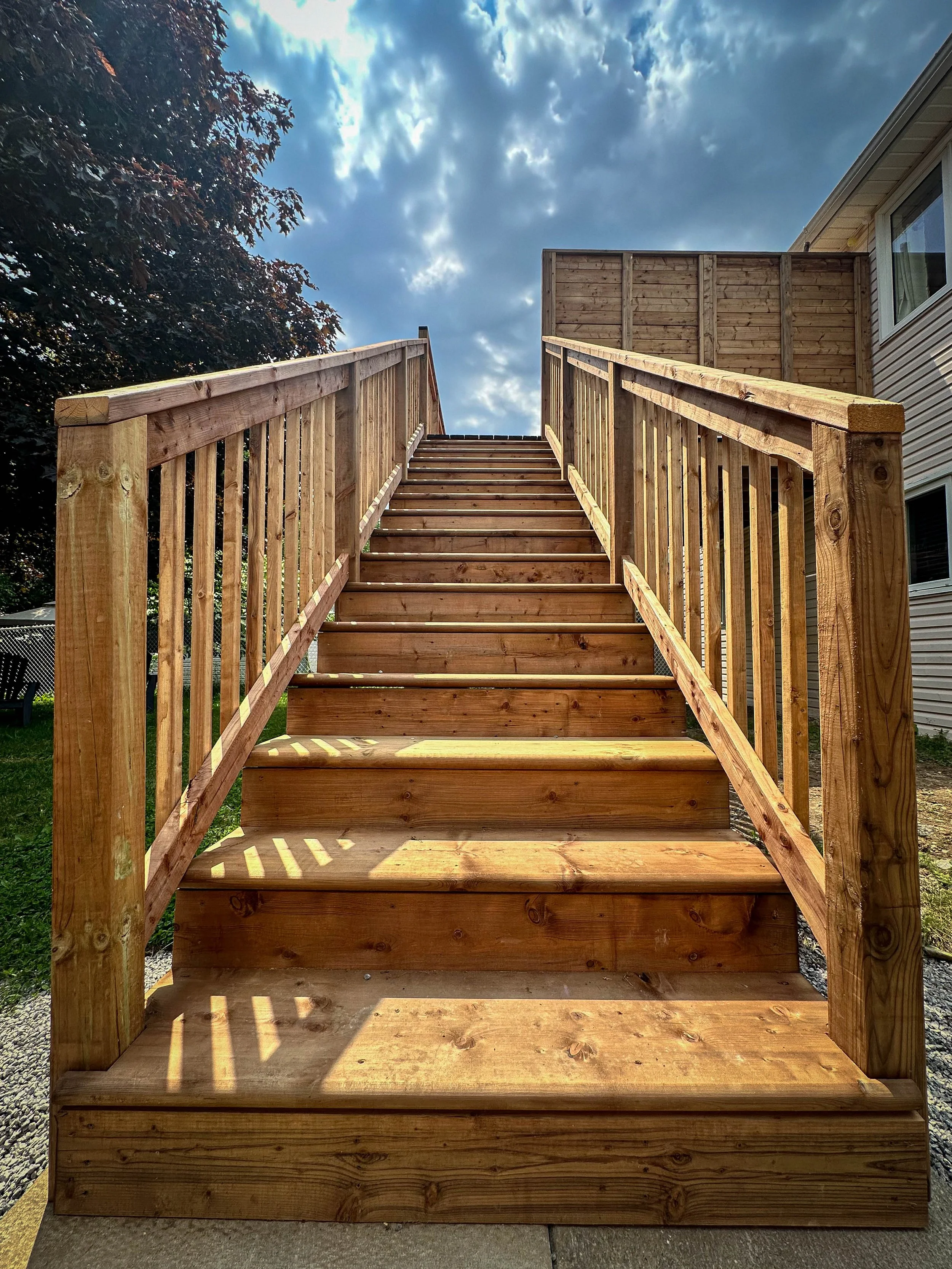 Wooden outdoor staircase leading up to a deck with a wooden railing on each side, outdoors under a partly cloudy sky.