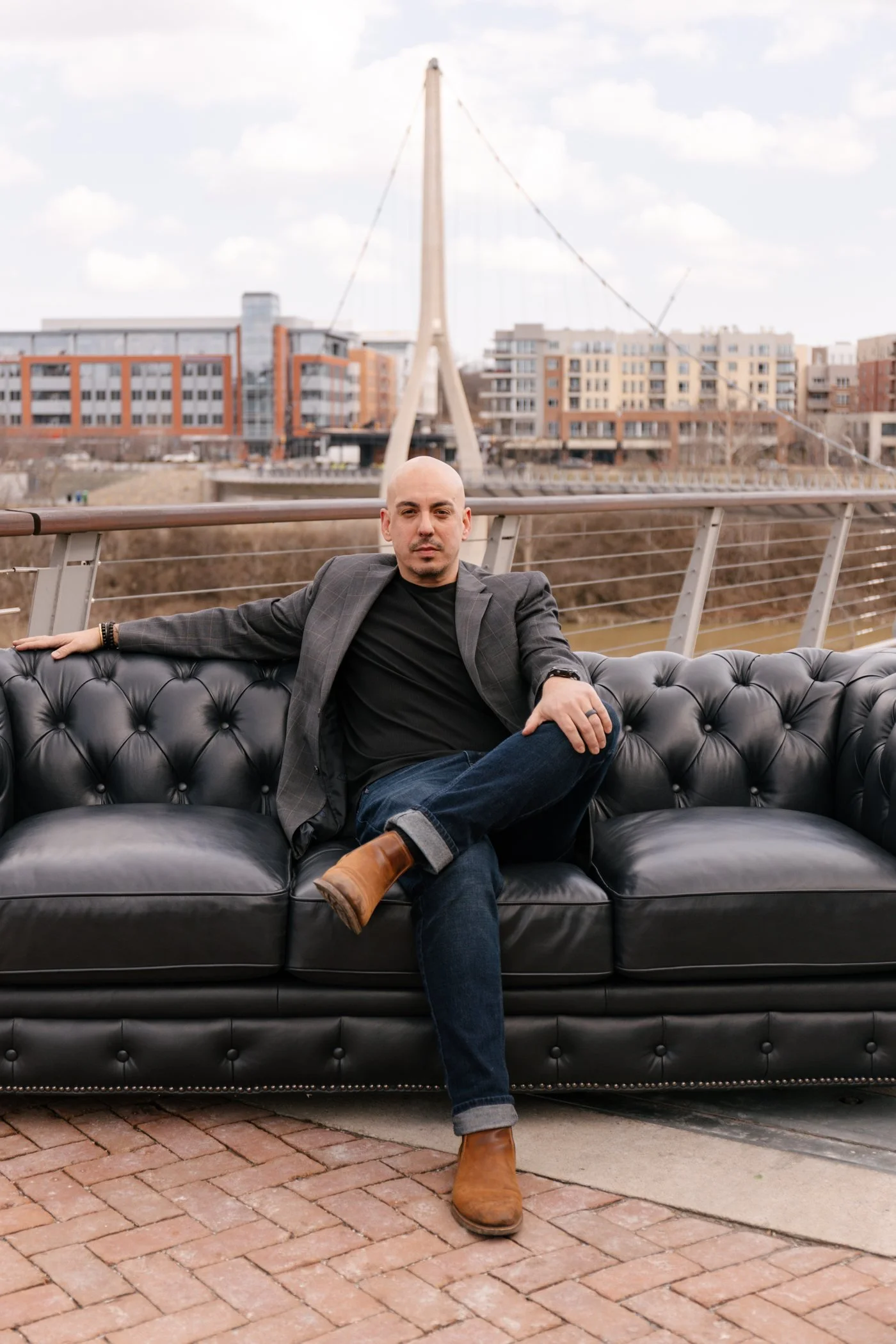 Yoshi Khosrowshahi seated on a black leather Chesterfield sofa outdoors, a cable-stayed pedestrian bridge and city buildings behind him