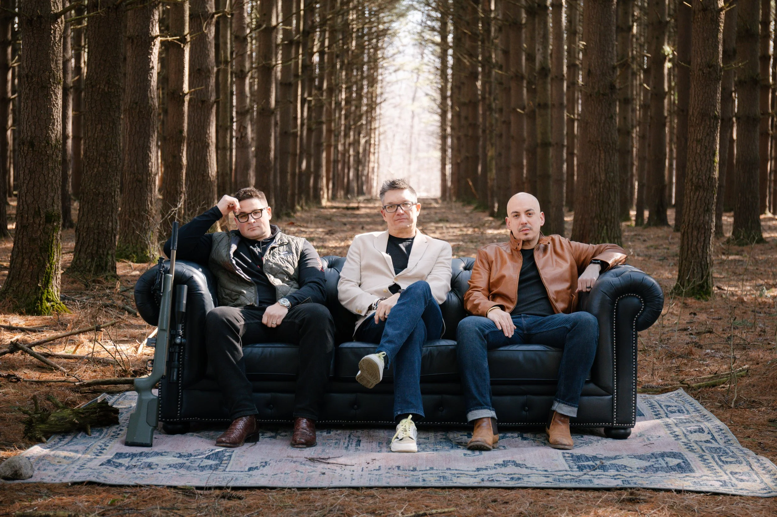 Three men sitting on a black couch in a forest, with tall trees surrounding them and sunlight filtering through the branches.