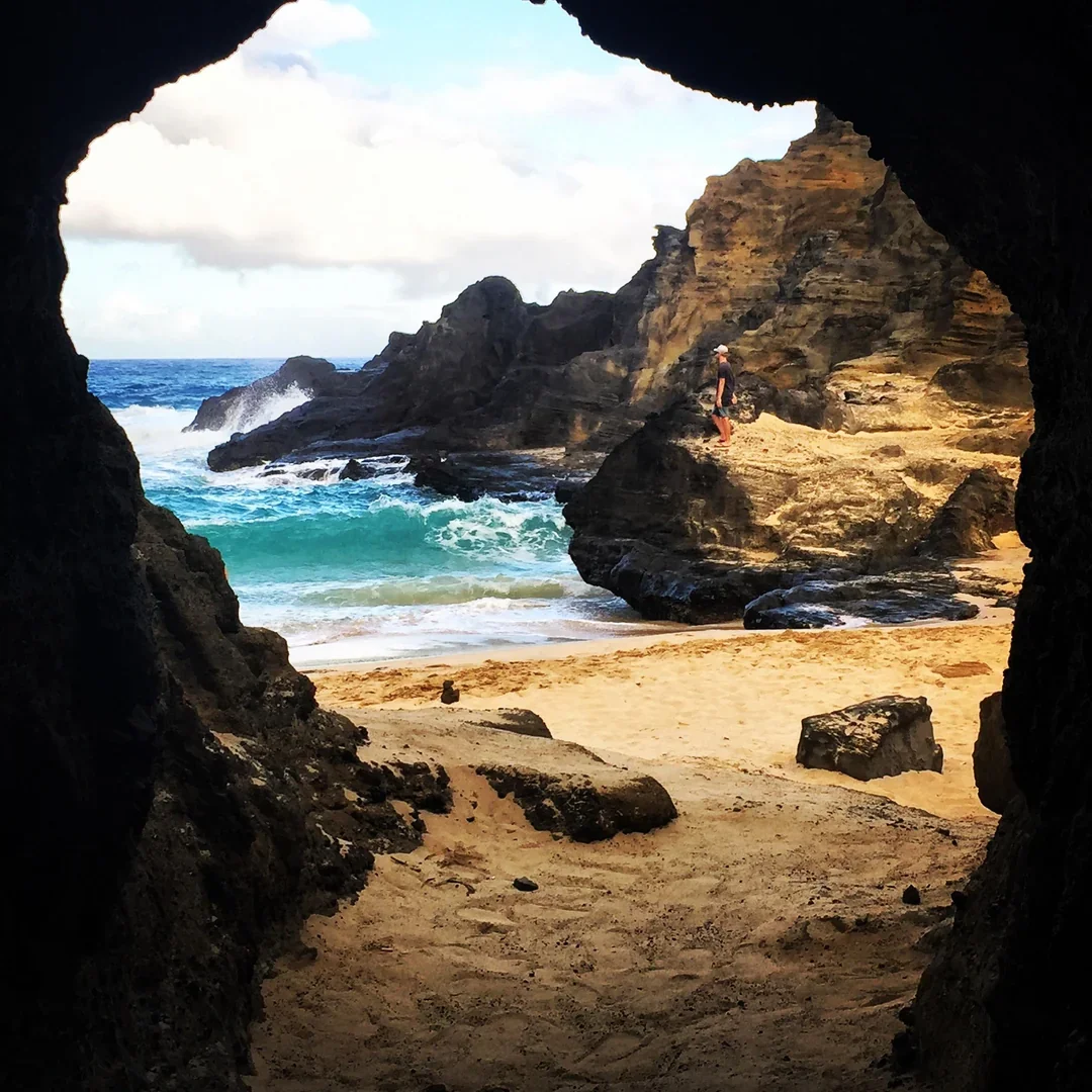View of a rocky beach seen through a cave opening, with a person standing on a rock formation near the shoreline and waves crashing in the ocean.