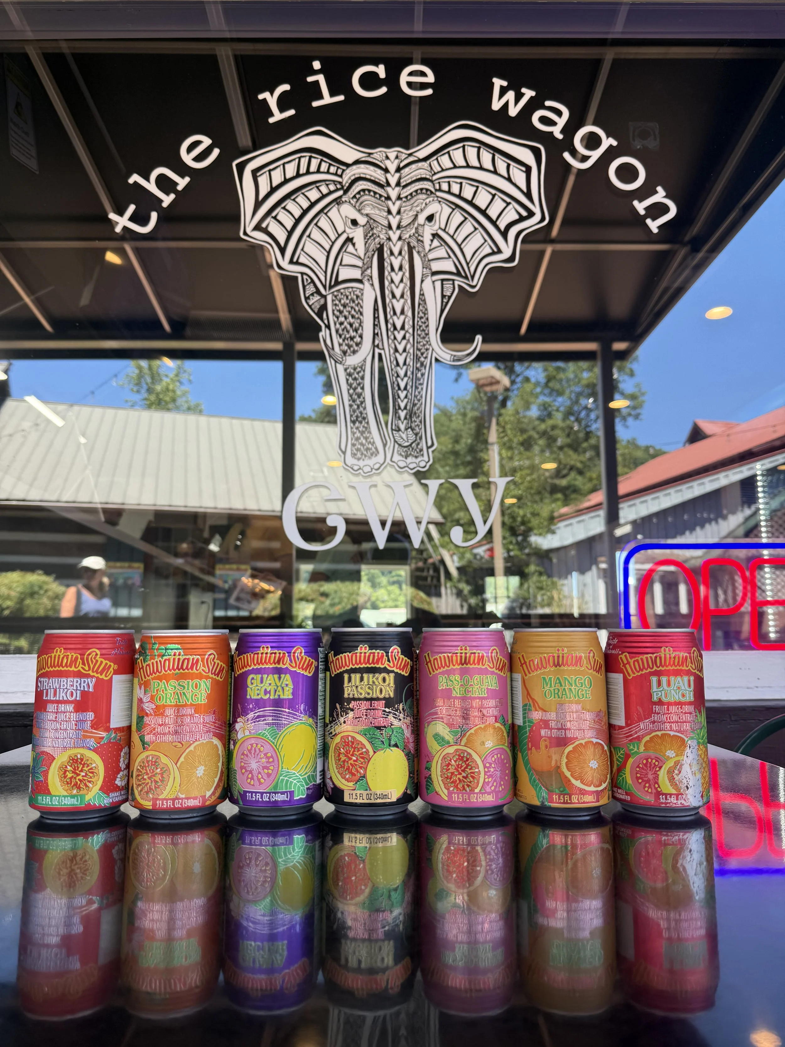 A row of Hawaiian Sun fruit juice cans in front of a store window with a graphic of an elephant with large ears, and the words 'the rice wagon' above it. The cans feature colorful illustrations of various fruits.