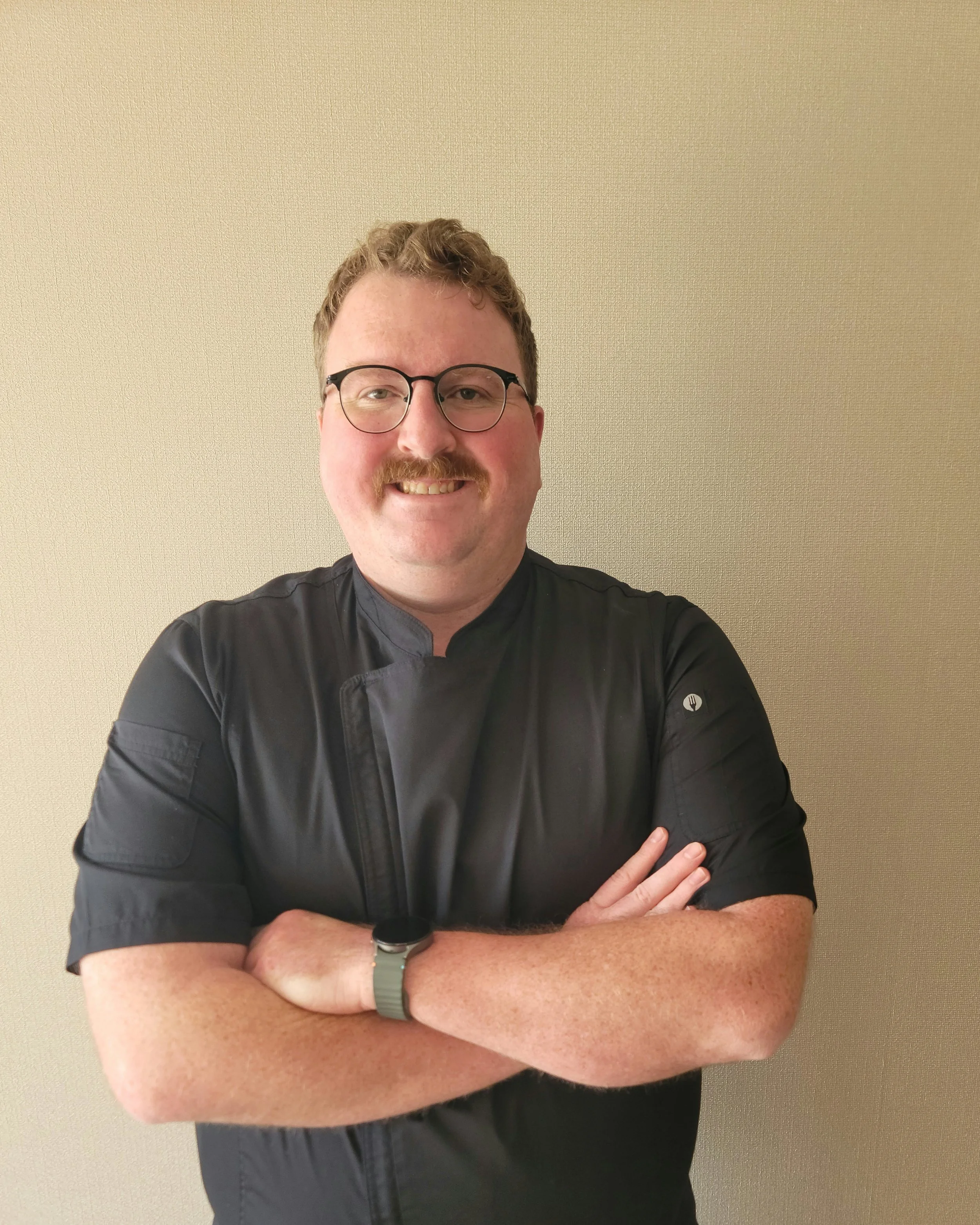 A man with short curly hair, glasses, and a mustache smiling with arms crossed. He is wearing a black/grey uniform top with and standing against a plain beige wall.