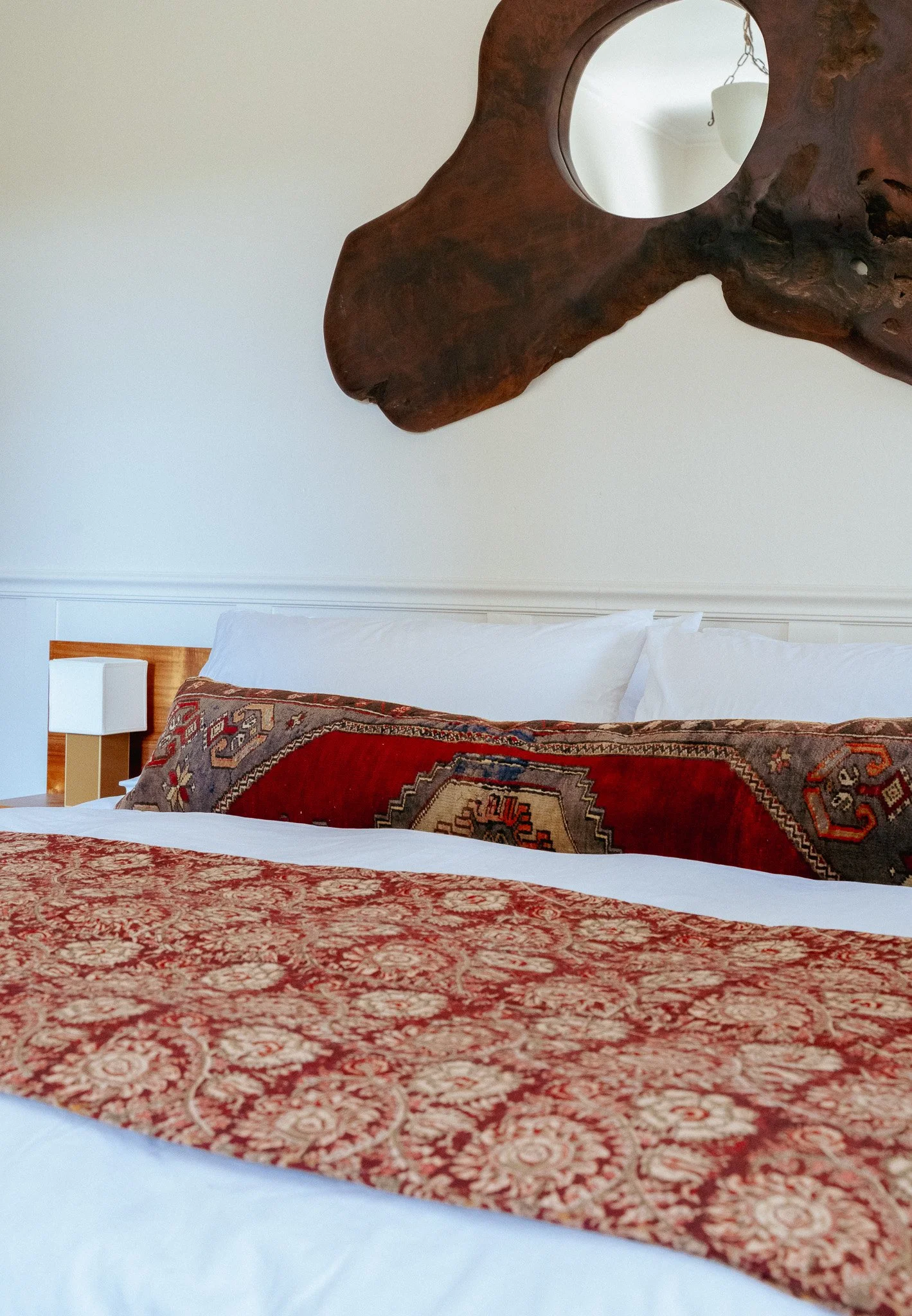 Interior of a bedroom showing a bed with a detailed red and beige patterned bedspread, white pillows, a wooden headboard, a bedside table with a lamp, and a decorative wooden wall hanging with a circular mirror.
