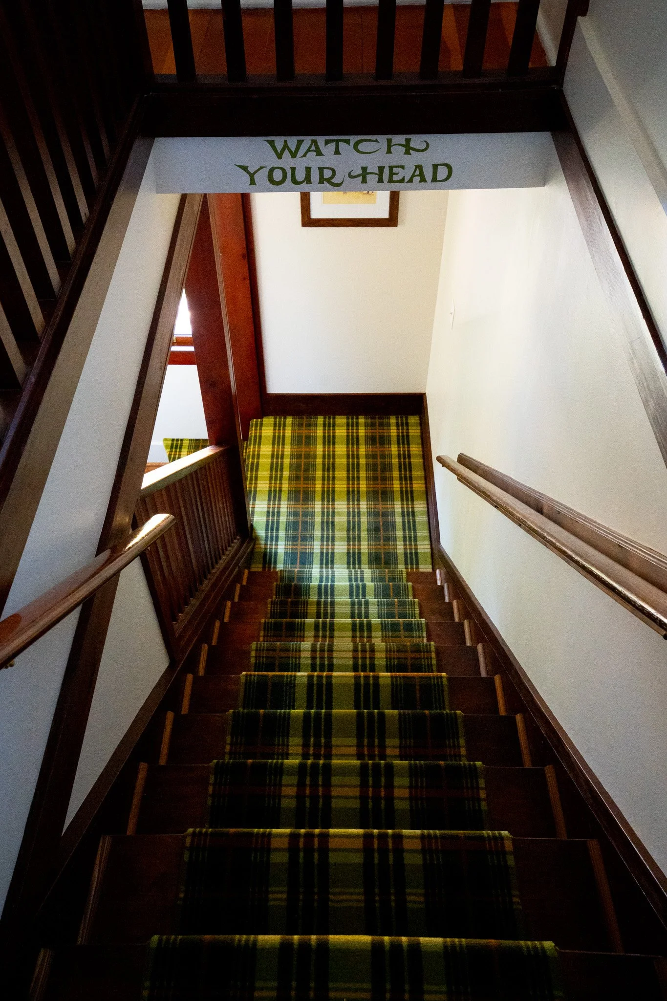 The photo shows a staircase with a patterned green, yellow, and black runner. There are wooden railings on both sides, and a sign hanging from the ceiling that says "WATCH YOUR HEAD" in green letters.