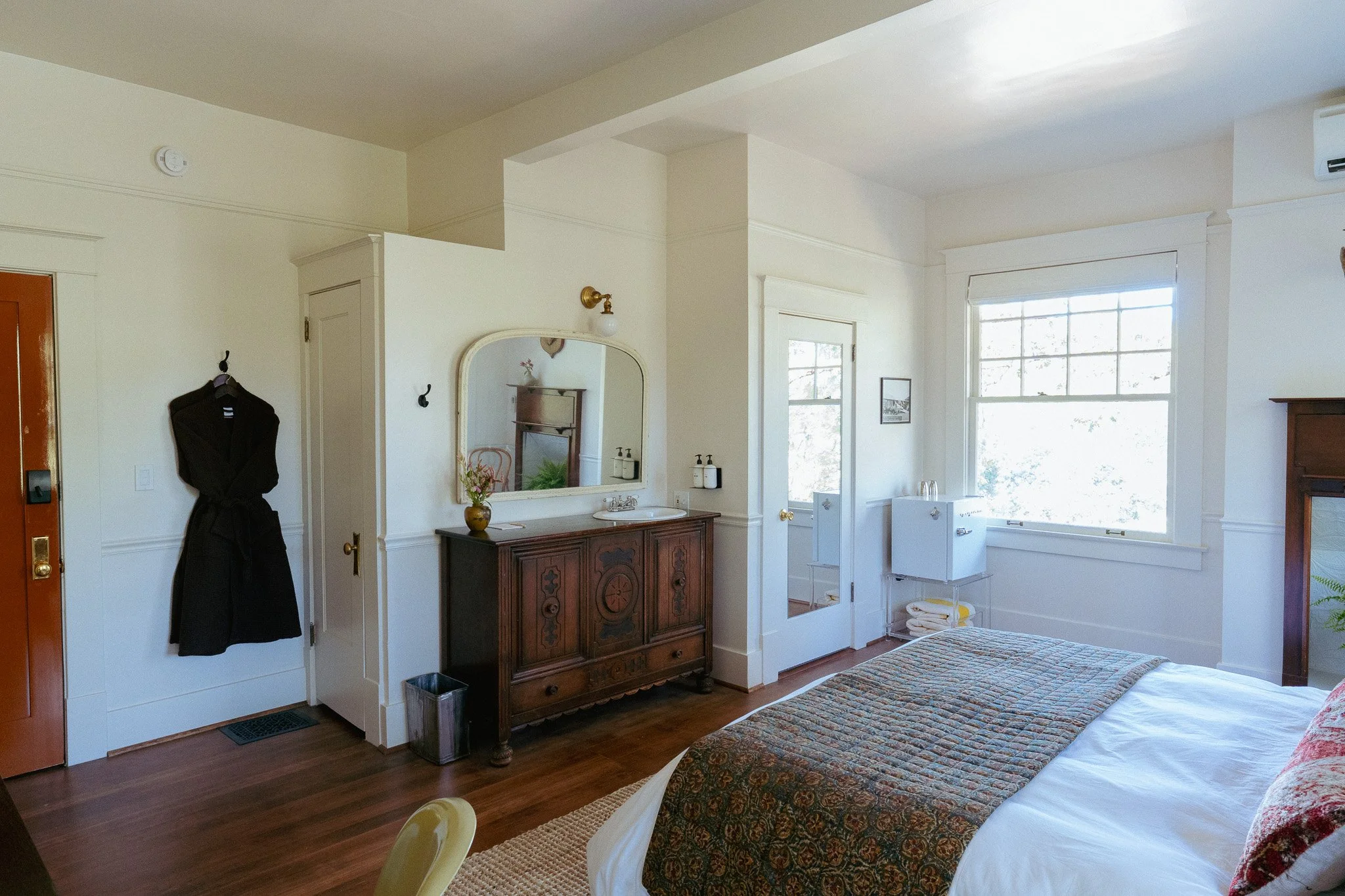 A cozy bedroom with a bed covered in a patterned quilt, a wooden dresser with decorative carvings, a large window letting in natural light, a mirror on the wall, and various small furniture and decor items.