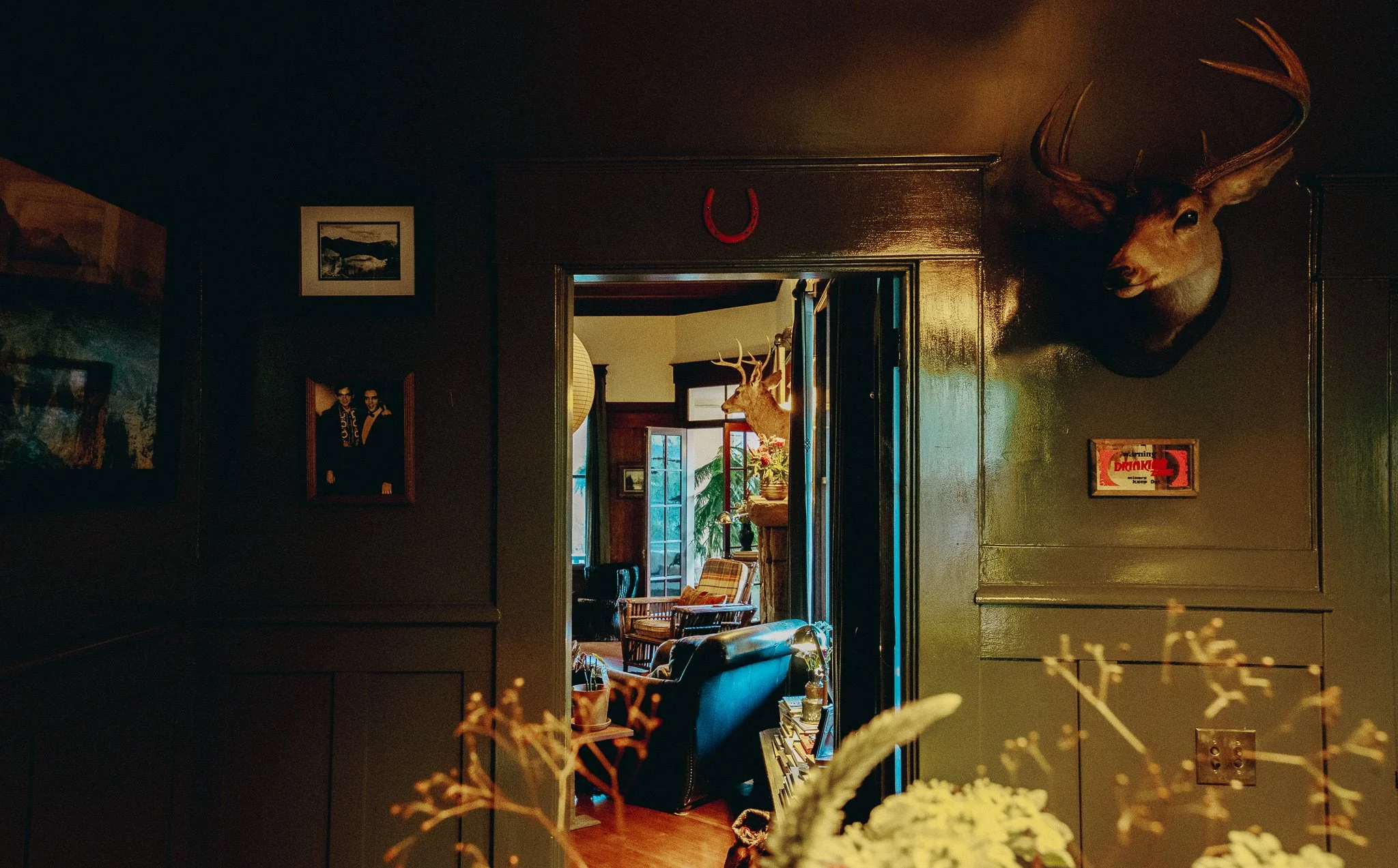 A dimly lit room with dark walls and mounted deer and elk heads on the wall. A doorway reveals another room with large windows, a plant, and furniture, including a wooden inclined chair.