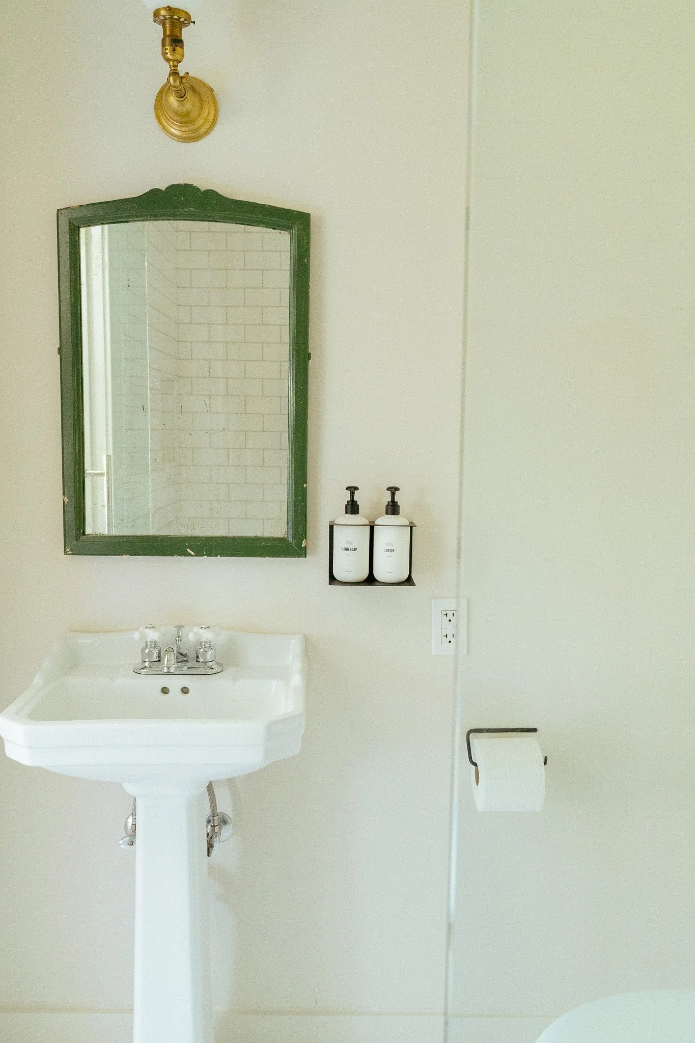 Bathroom with a white pedestal sink, a mirror with a green frame, two bottles of hand soap and lotion, a wall-mounted light fixture, a toilet paper holder with a roll of toilet paper, and a white wall with a power outlet.