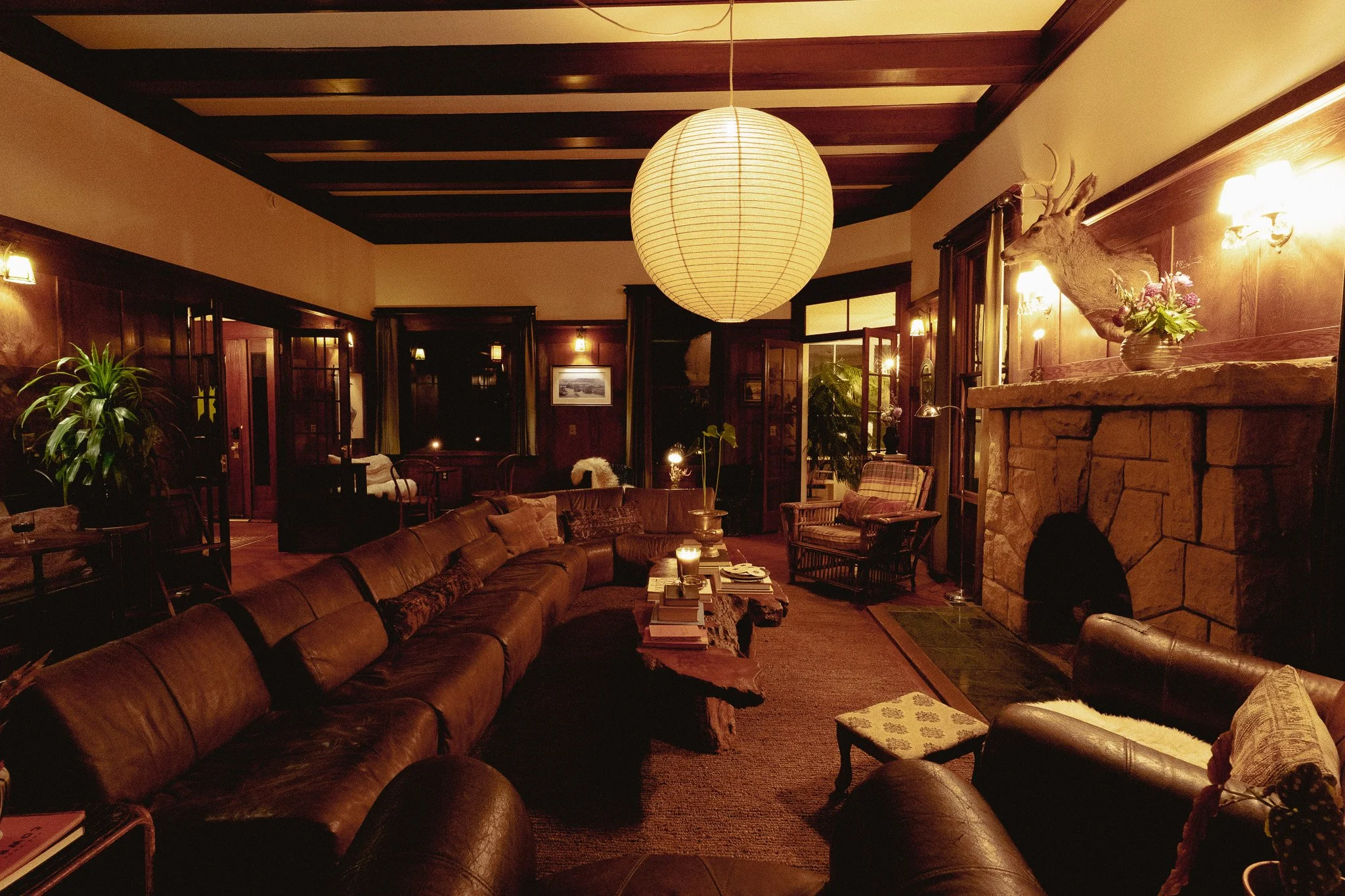 A cozy living room with dark wooden walls and ceiling beams, a large paper lantern hanging from the ceiling, a stone fireplace with a deer head mounted on it, potted plants, a leather sofa, and various chairs and tables illuminated by warm lighting.
