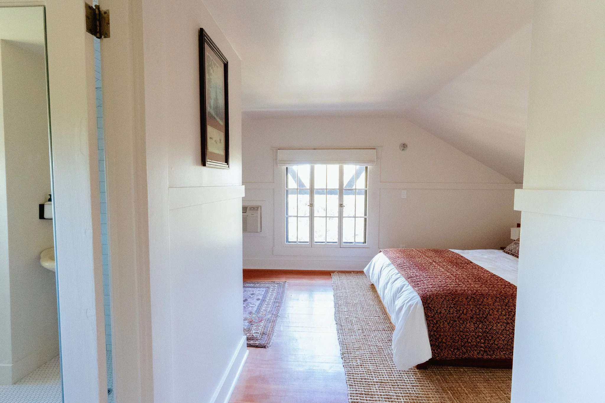 A bedroom with a large window, a bed with a red patterned blanket, a patterned rug, and a framed picture on the wall.