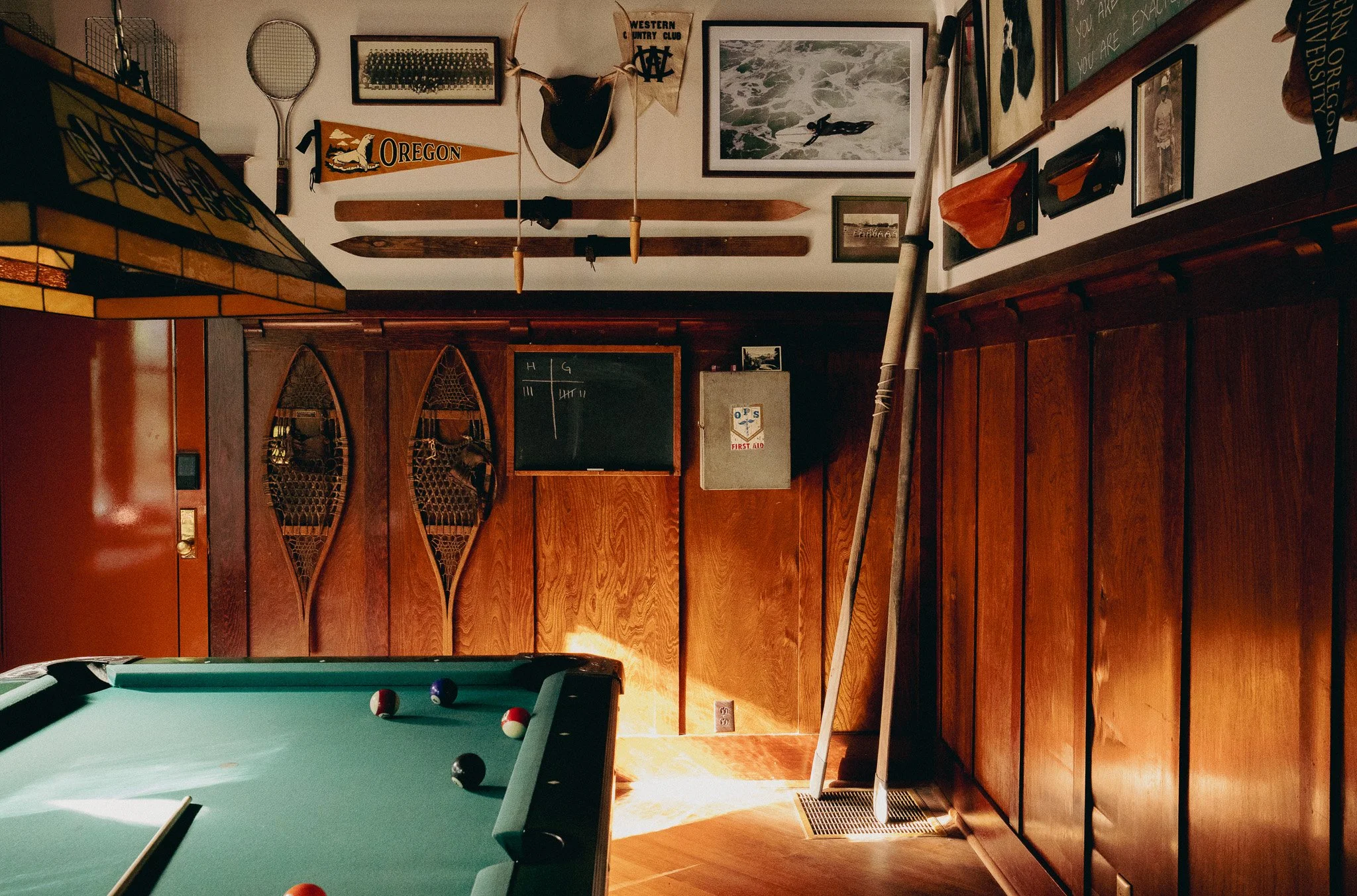 A view of a game room with a pool table in the foreground and a wall decorated with sports memorabilia, framed pictures, wooden paddles, a tennis racket, antlers, and flags, all with wood paneling below.