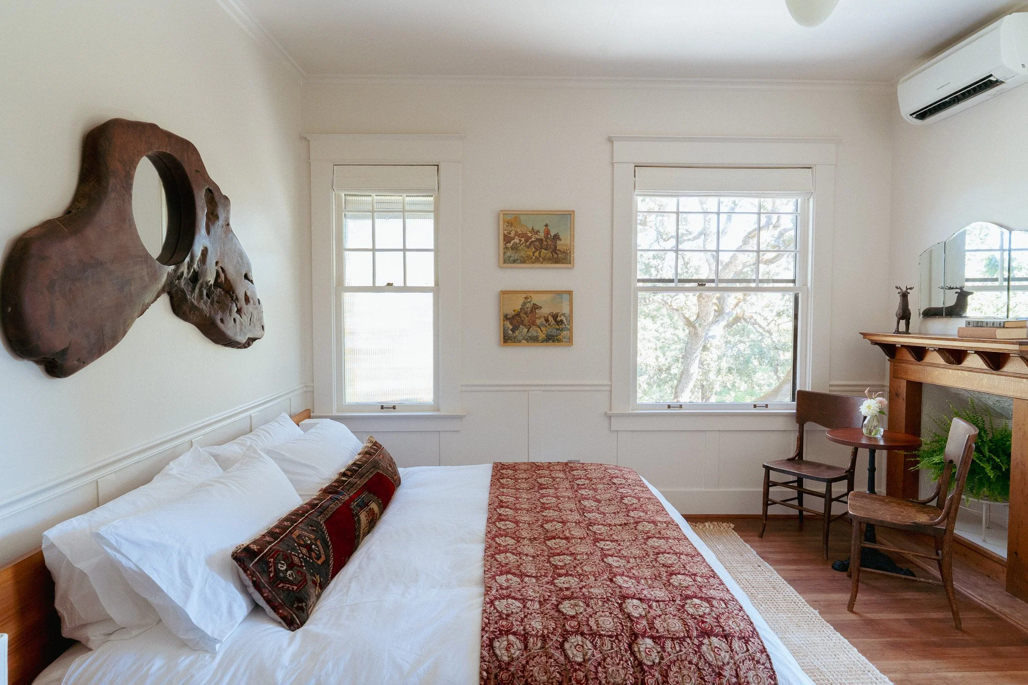 A cozy bedroom with white walls and large windows showing trees outside. The bed has white linens, patterned pillows, and a red patterned runner. There is a wooden mantle with decorative items, including two small deer sculptures, and framed artwork on the wall.