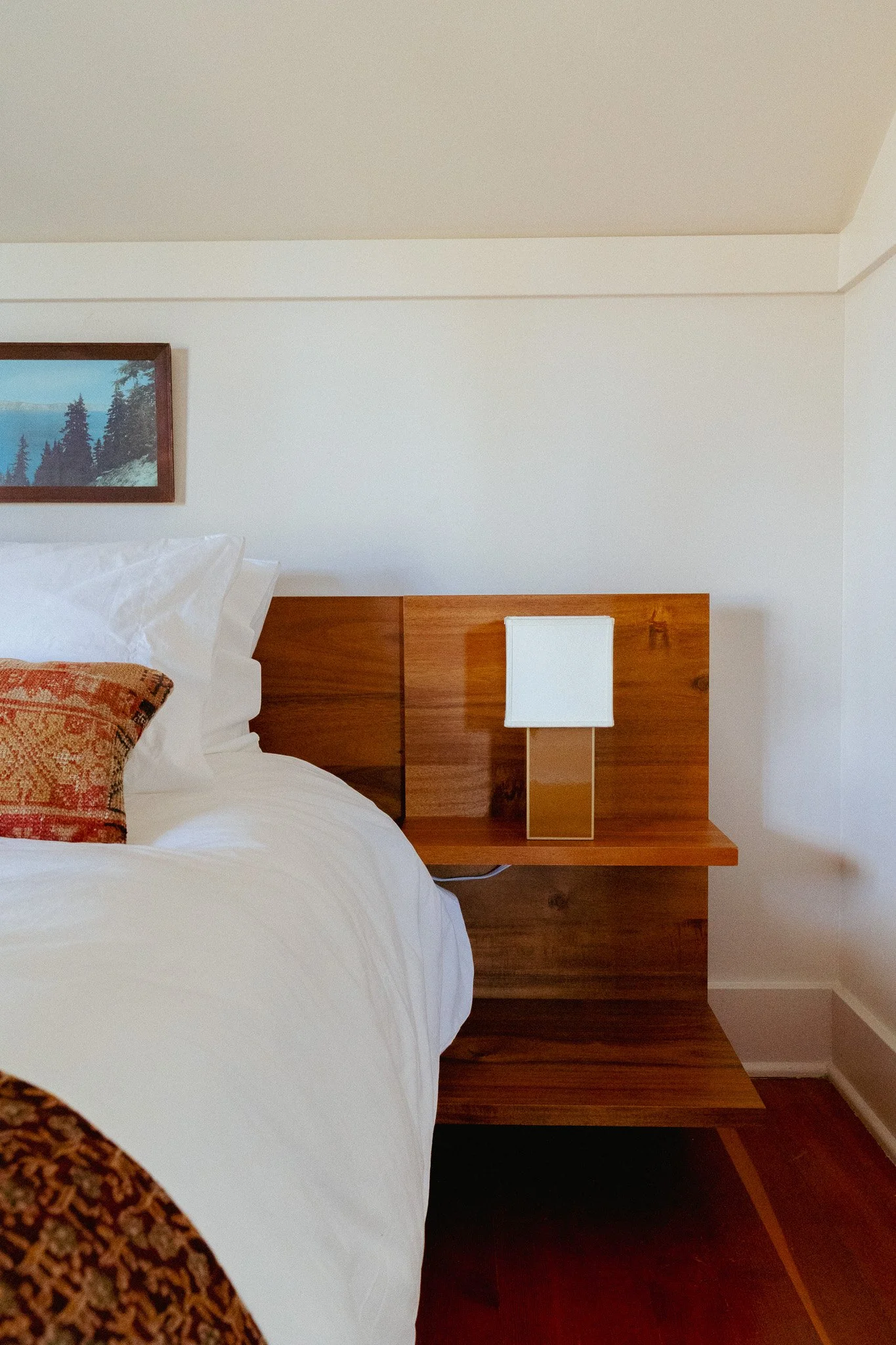 A bedroom with a white bed, decorative pillows, wooden headboard, and modern bedside lamp. A framed landscape painting hangs above the bed, and beige walls with crown molding are visible.