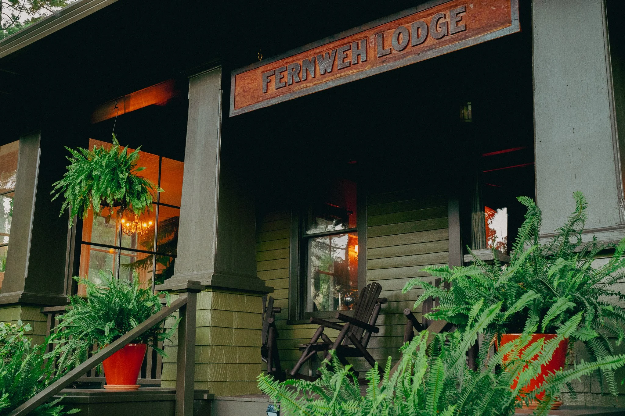 Exterior of Fernweh Lodge with wooden sign, large windows, and surrounding green plants on a porch.