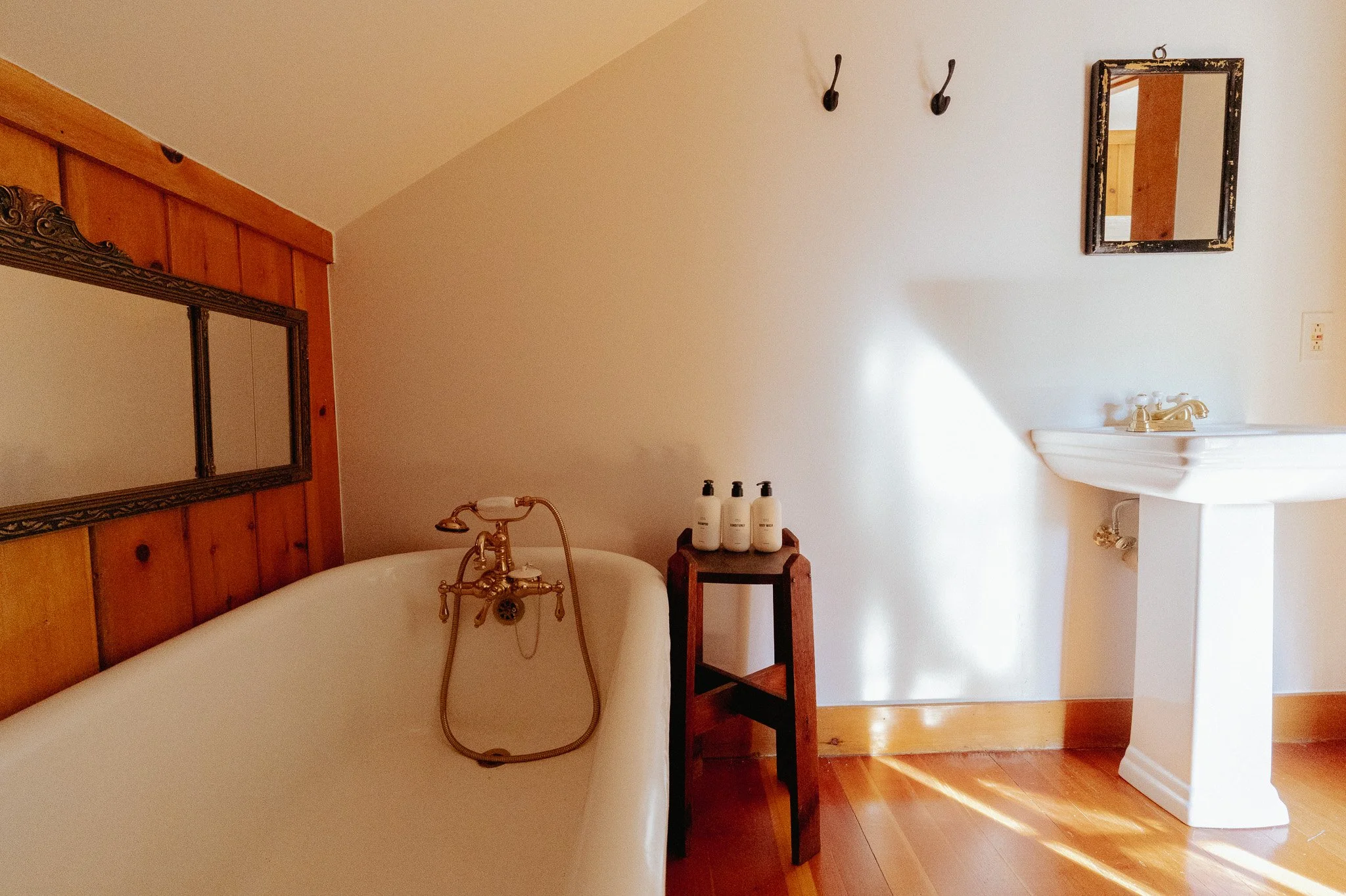 A bathroom with a vintage clawfoot bathtub featuring brass fixtures, framed mirrors, hooks on the wall, and bottles of toiletries on a small shelf.