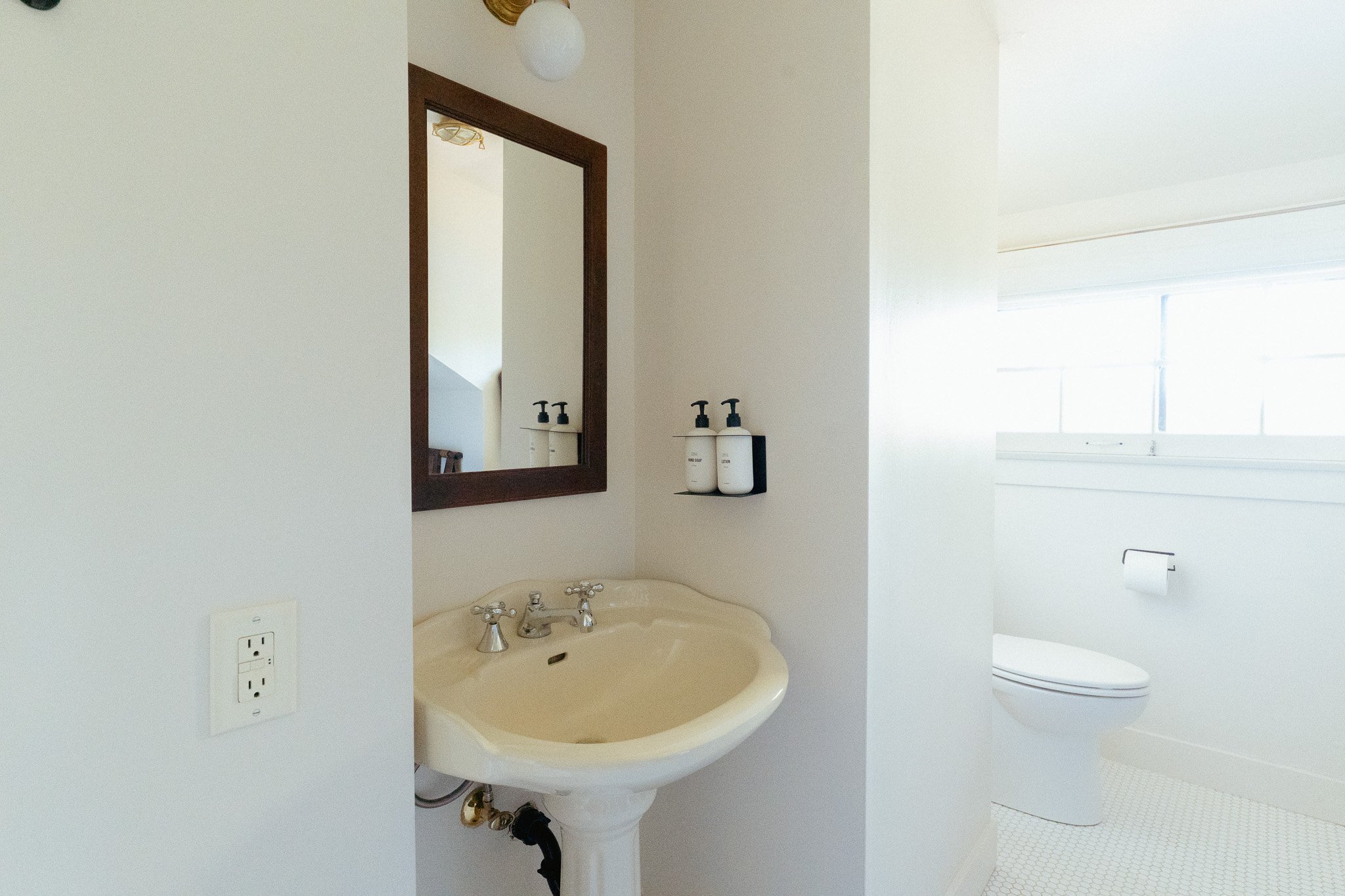 A bathroom with a vintage sink, three soap dispensers on a black tray, a mirror, a window with blinds, and a toilet.