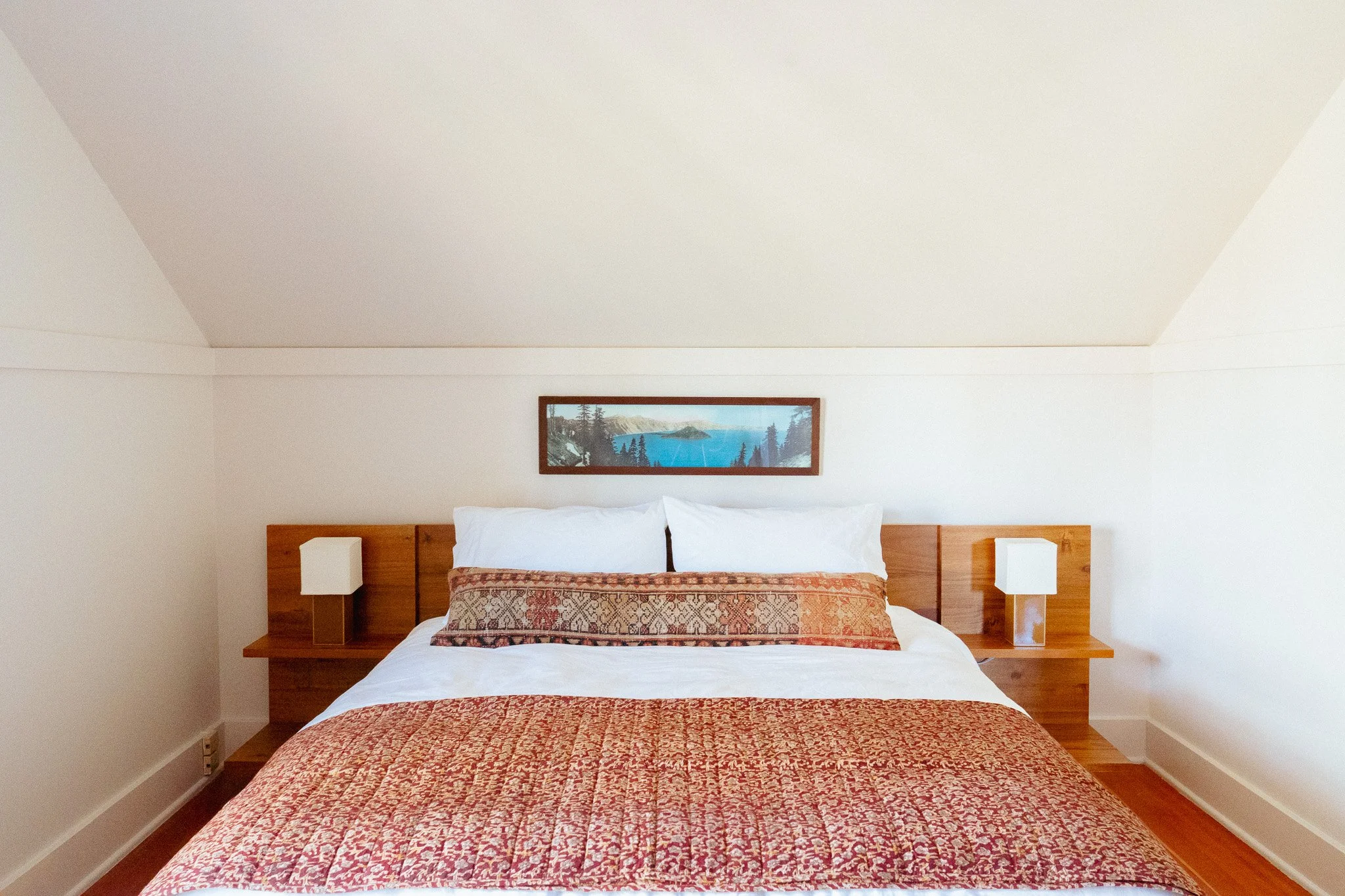 A tidy bedroom with a large bed covered with a red patterned bedspread and a white top sheet, flanked by two wooden nightstands with white lamps, hung with a framed landscape painting of a lake surrounded by trees and mountains.