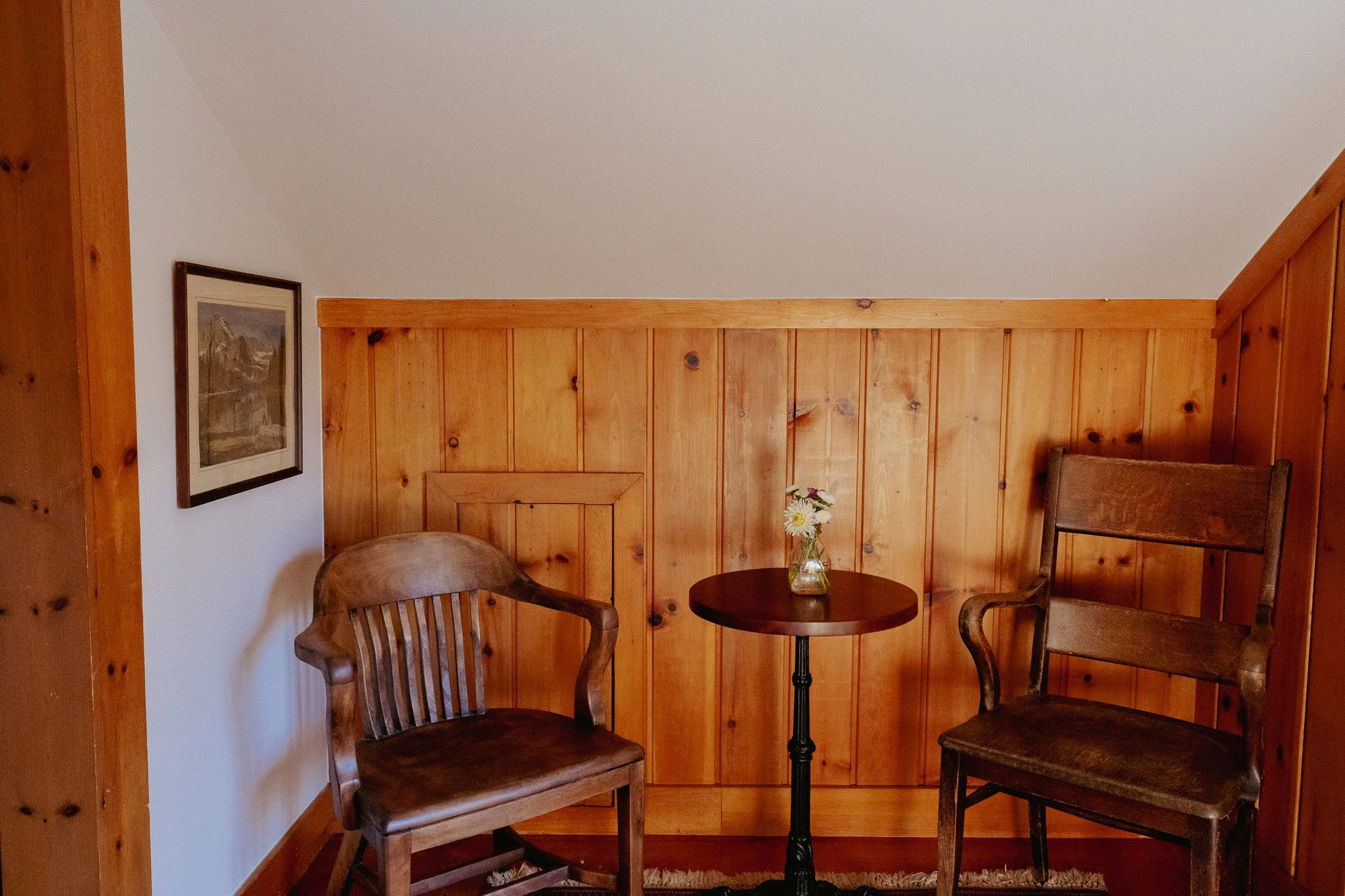 A cozy corner of a room with wooden paneled walls, featuring two wooden chairs around a small round table with a vase of flowers. There is a framed picture on the white wall to the left.