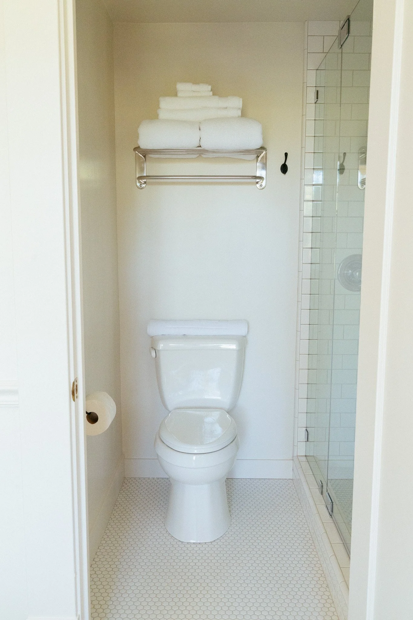 Bathroom with a toilet, toilet paper roll, towels on a shelf, and a glass shower door with tiled interior.