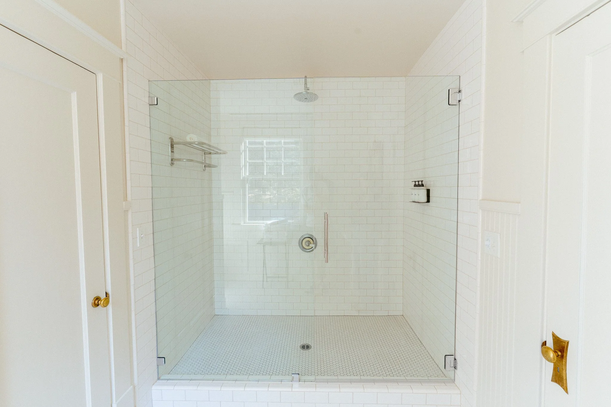 A modern walk-in shower with clear glass walls, white subway tile on the walls, a rain showerhead on the ceiling, a small shelf with soap, and a drain on the floor.