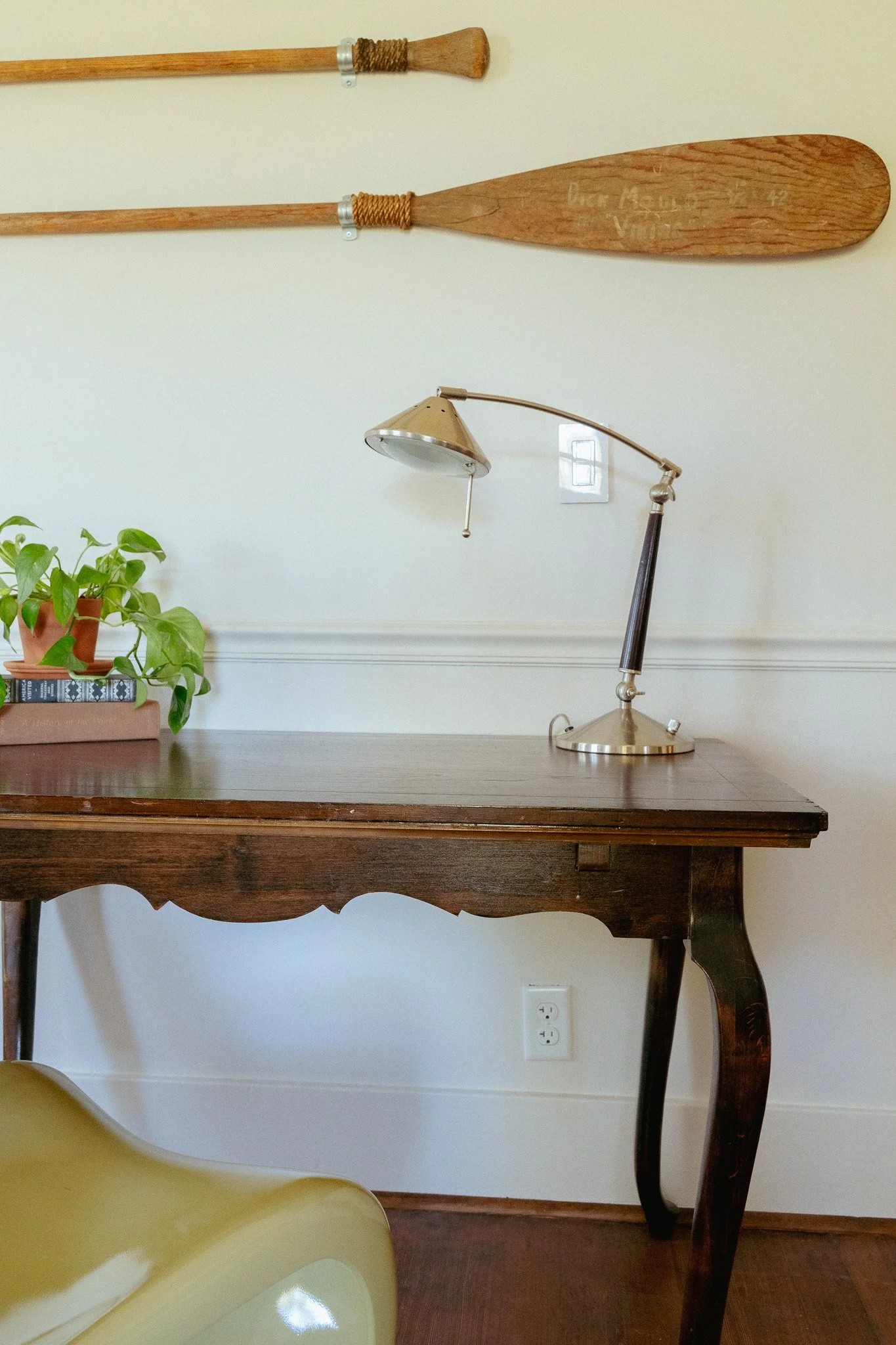 A wooden desk with a yellow chair at the bottom left corner. On the desk, there is a potted plant and a book. The background shows a white wall with a power outlet, a vintage desk lamp, and two wooden oars mounted horizontally on the wall.