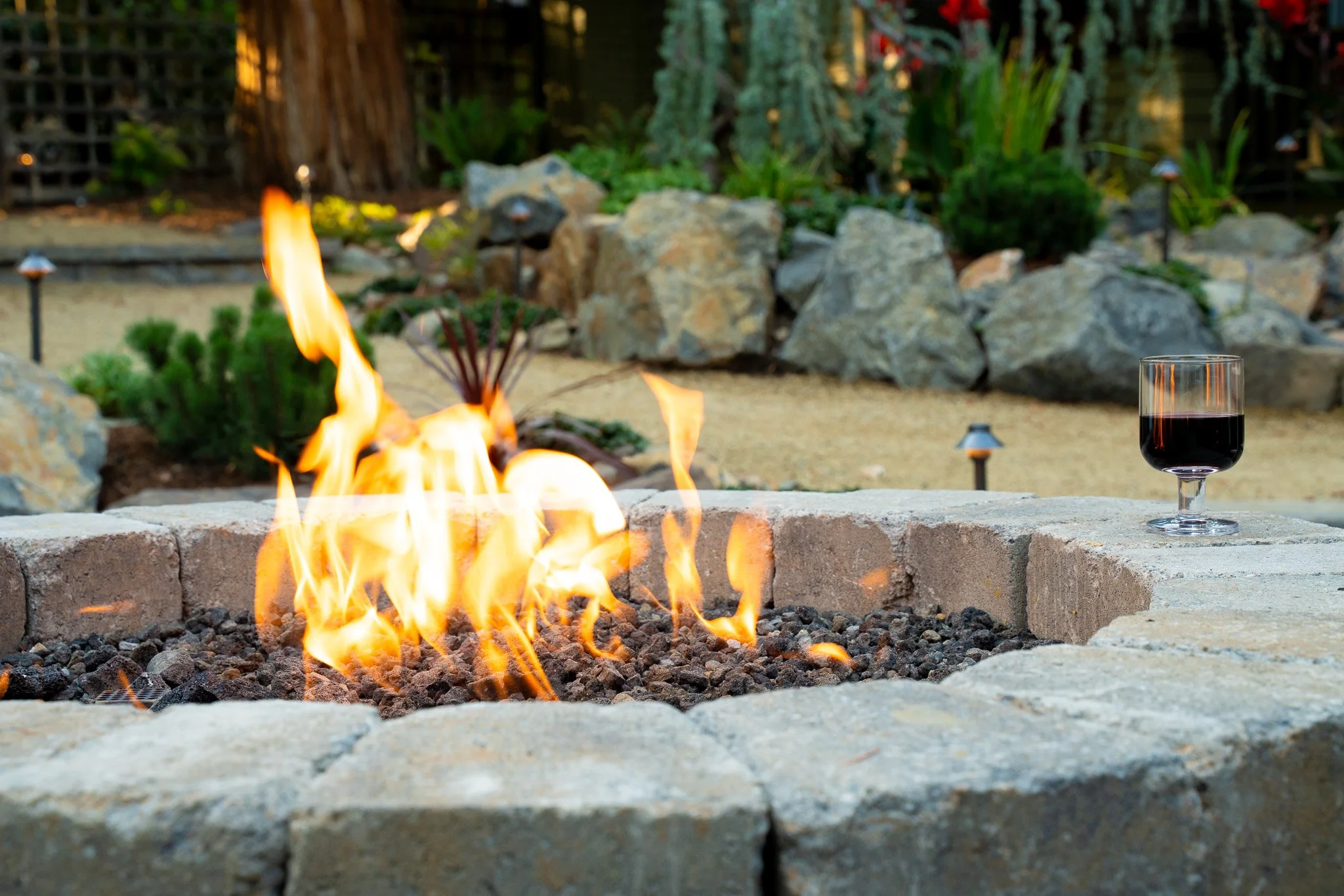 A backyard patio with a fire pit, a glass of red wine, garden rocks, plants, and small pathway lights, during the evening.