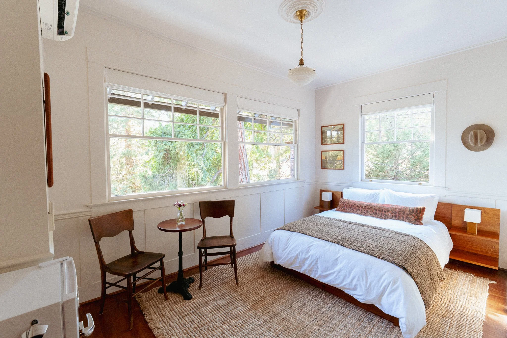 A bedroom with a white ceiling and walls, three large windows with white blinds showing greenery outside, a bed with white bedding and a decorative pillow, a wooden nightstand with a lamp, three framed pictures on the wall, and two chairs around a small round table with a vase of flowers. A beige rug covers the wooden floor, and a hat hangs on the wall.