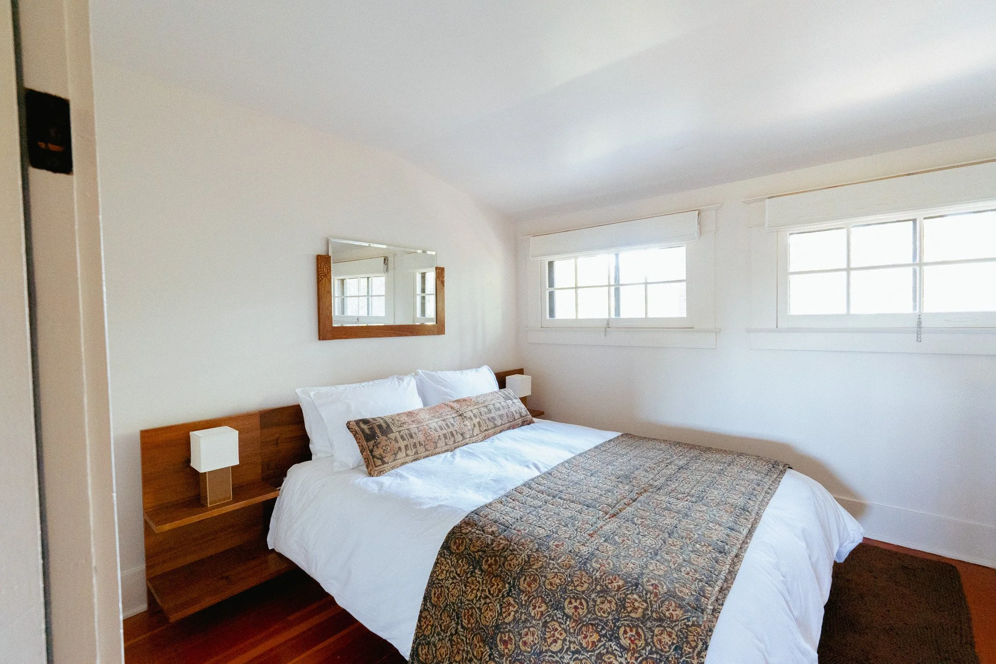 A bedroom with white walls, a wooden bed with white bedding, decorative pillows, and a quilt. There are two windows with sunlight coming through and a mirror on the wall above the bed.