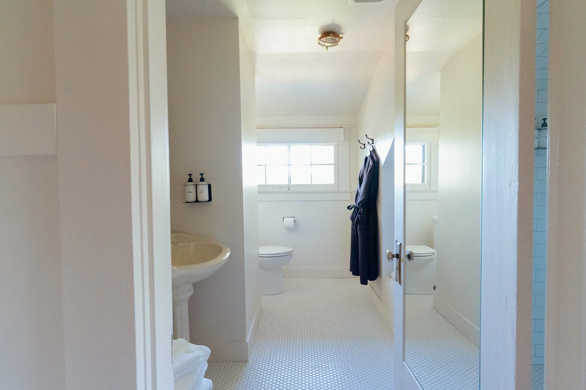 View of a bathroom seen through an open door. It has a white wall with a small window, a toilet, a bathtub, and a black towel hanging on a hook. Sunlight fills the room from the window.