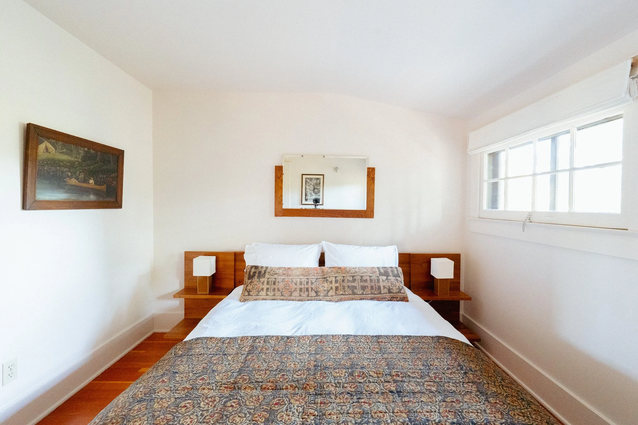 A bedroom with a white bed, two white nightstands, and wall-mounted lamps. There are two framed pictures on the wall, one above the bed and a mirror above the headboard. A large window with white blinds is on the right side.