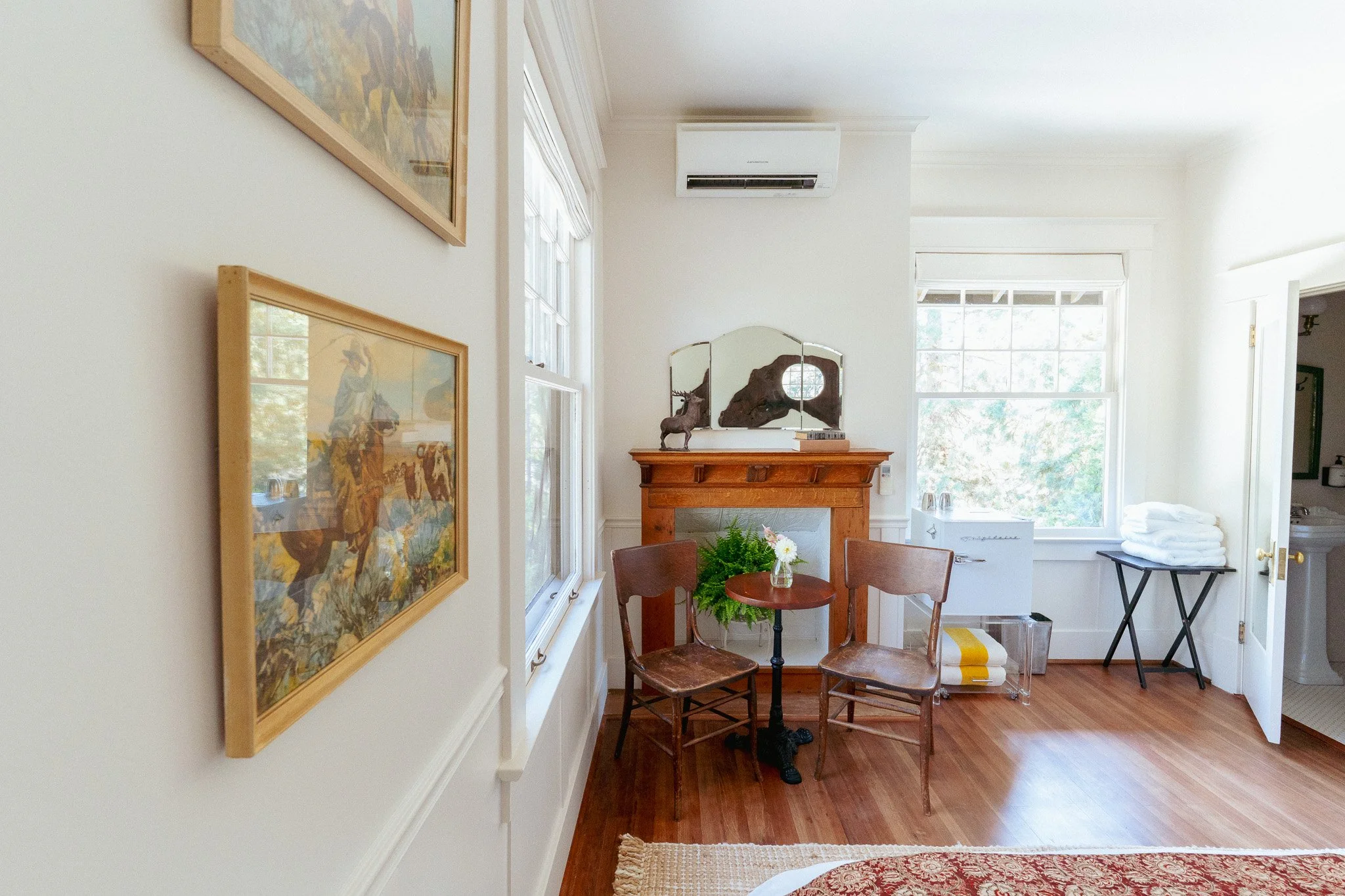 A cozy living room with a small round table and two wooden chairs in front of a fireplace decorated with three mirrors and a deer figurine. Large windows allow natural light to illuminate the space, which also features framed artwork on the walls and an air conditioner mounted above the fireplace.