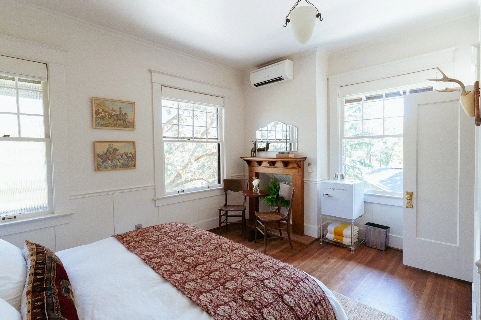 A cozy bedroom with white walls and large windows, featuring a bed with a patterned red and gold bedspread, a small wooden table with chairs, a fireplace, and decorative items including paintings and a mounted deer head.