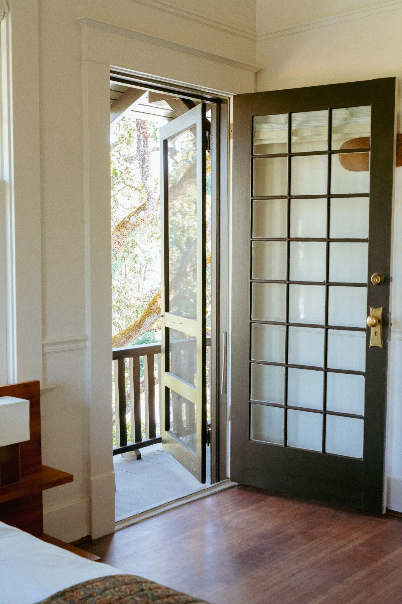 Open glass-paneled door leading to a small balcony with wooden railing, outside view of trees and sunlight.