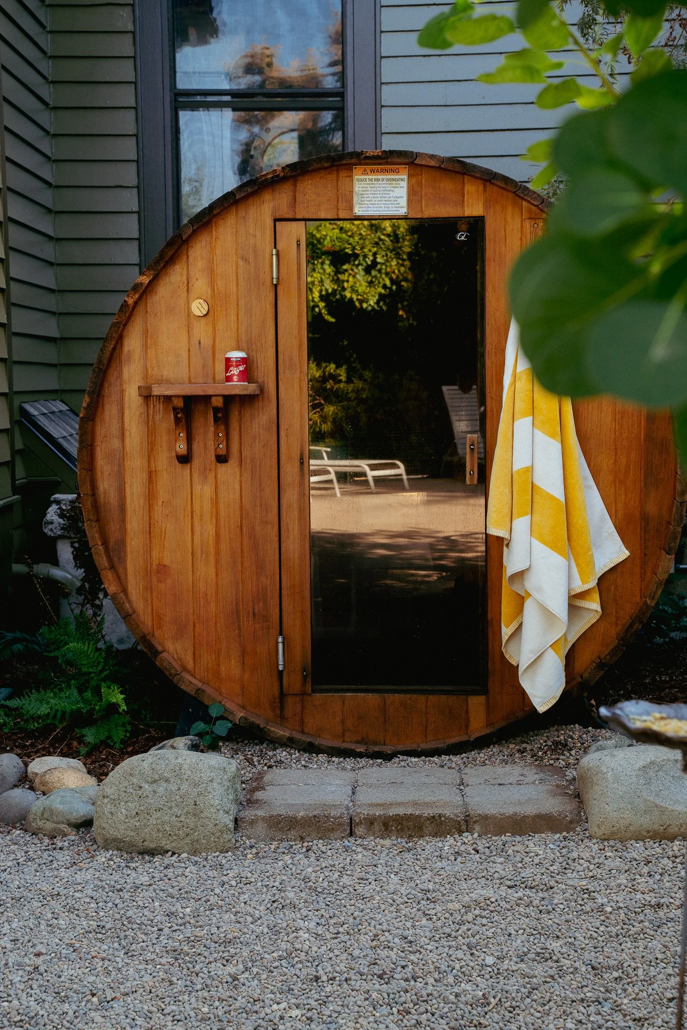 A wooden, barrel-shaped outdoor shower with a glass door, a mirror, a small shelf with a red can, a yellow and white towel hanging on the side, surrounded by rocks and greenery.