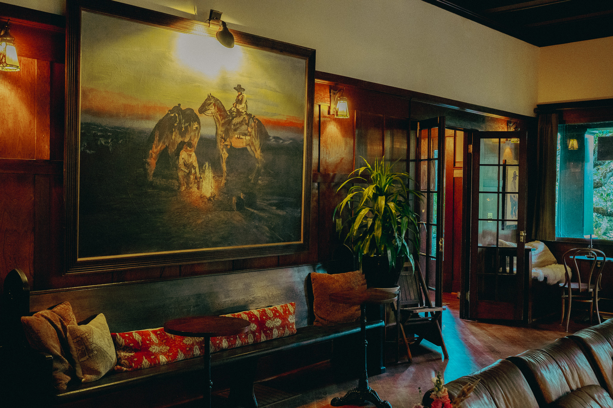 A cozy living room featuring a large painting of three cowboys on horses during sunset, wooden paneled walls, a plush floral sofa with cushions, hanging lamps, a houseplant, and open glass doors leading outside.