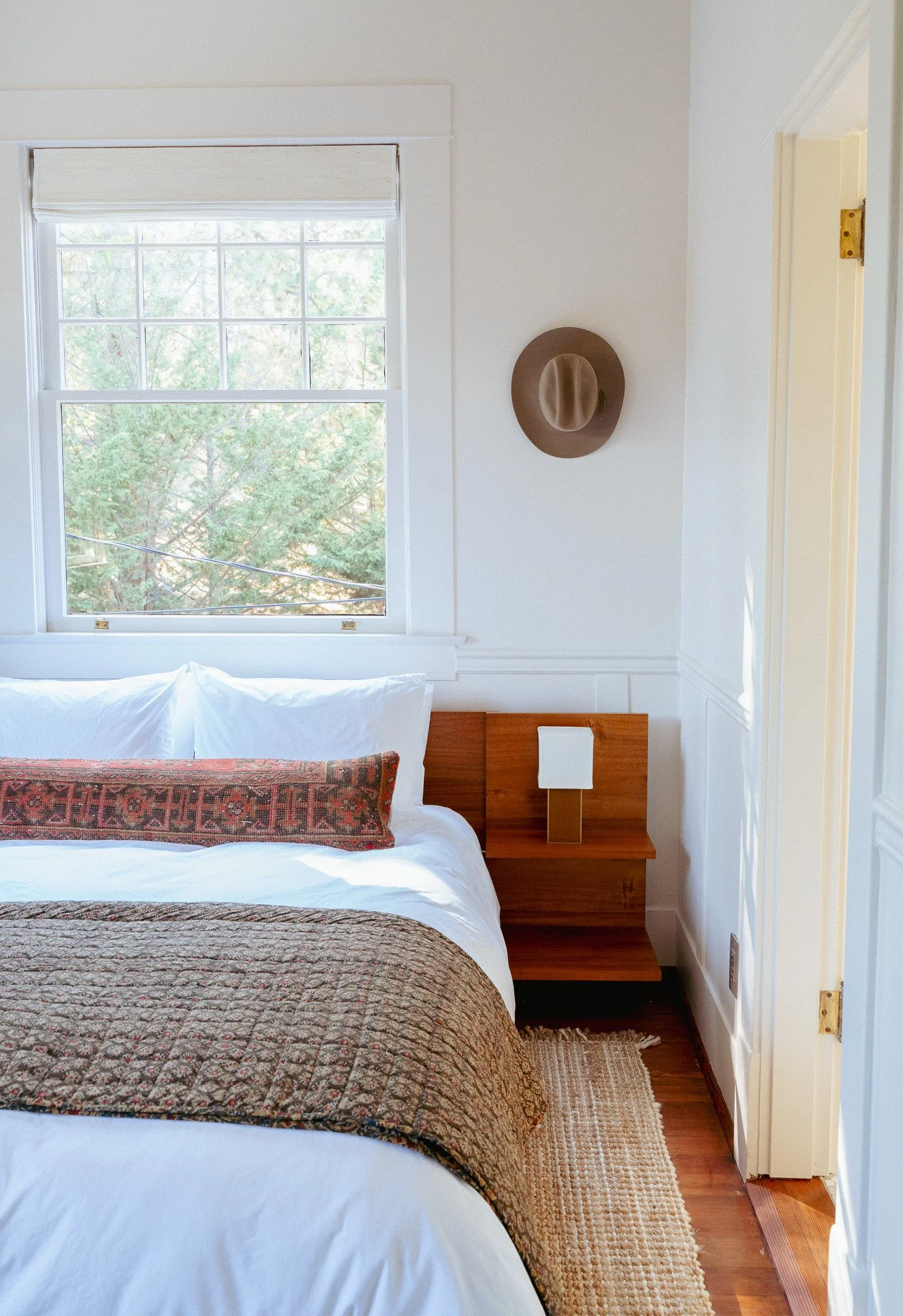A bedroom with a bed that has a patterned quilt and a decorative pillow. A window with a white blind shows green trees outside. A wooden nightstand holds a white lamp and a hat hangs on the wall.