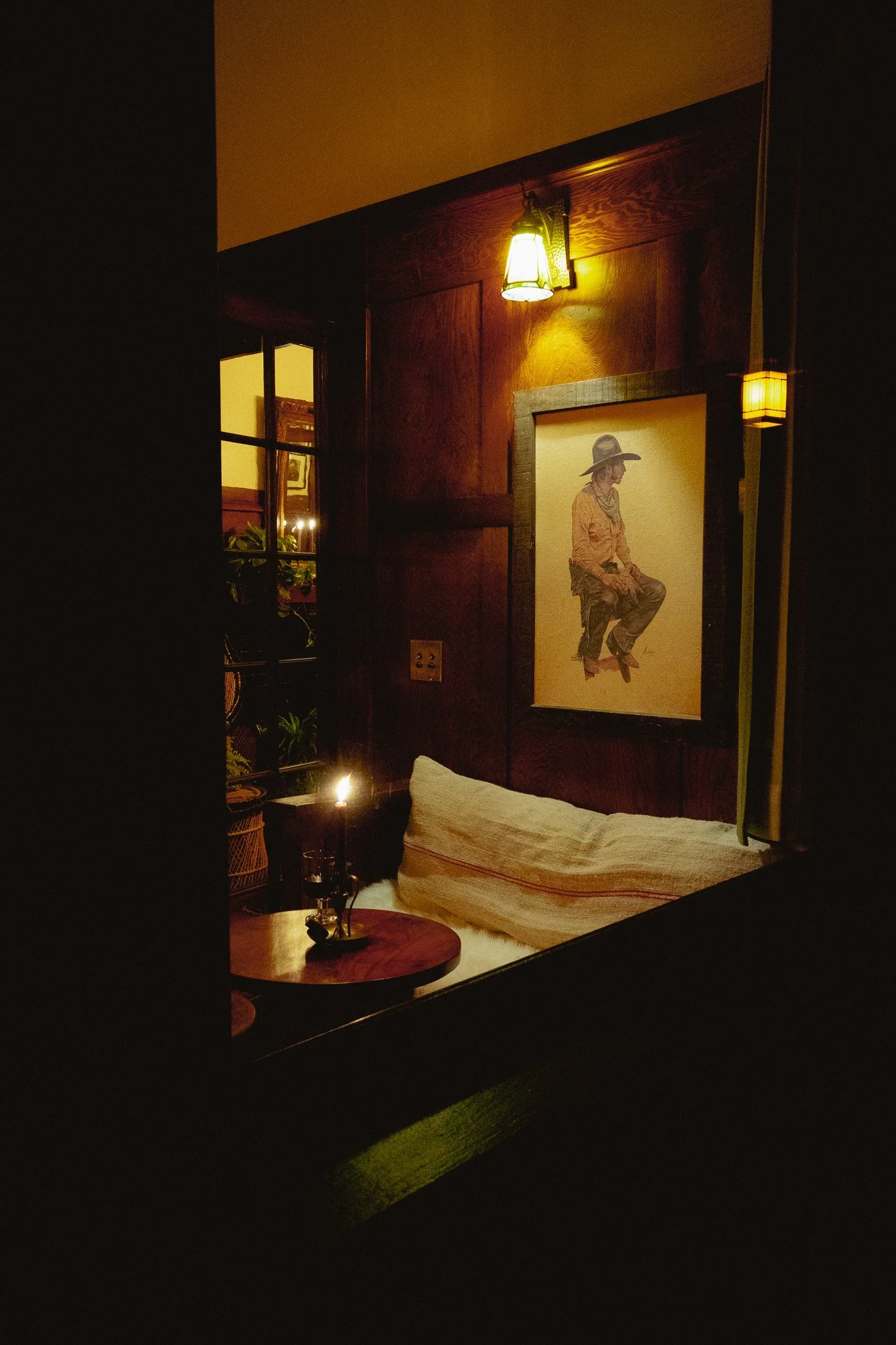 Dimly lit bedroom corner with wooden wall paneling, a framed artwork of a woman in a hat, a small round wooden side table with a candle, and a bed with a folded blanket and pillow.
