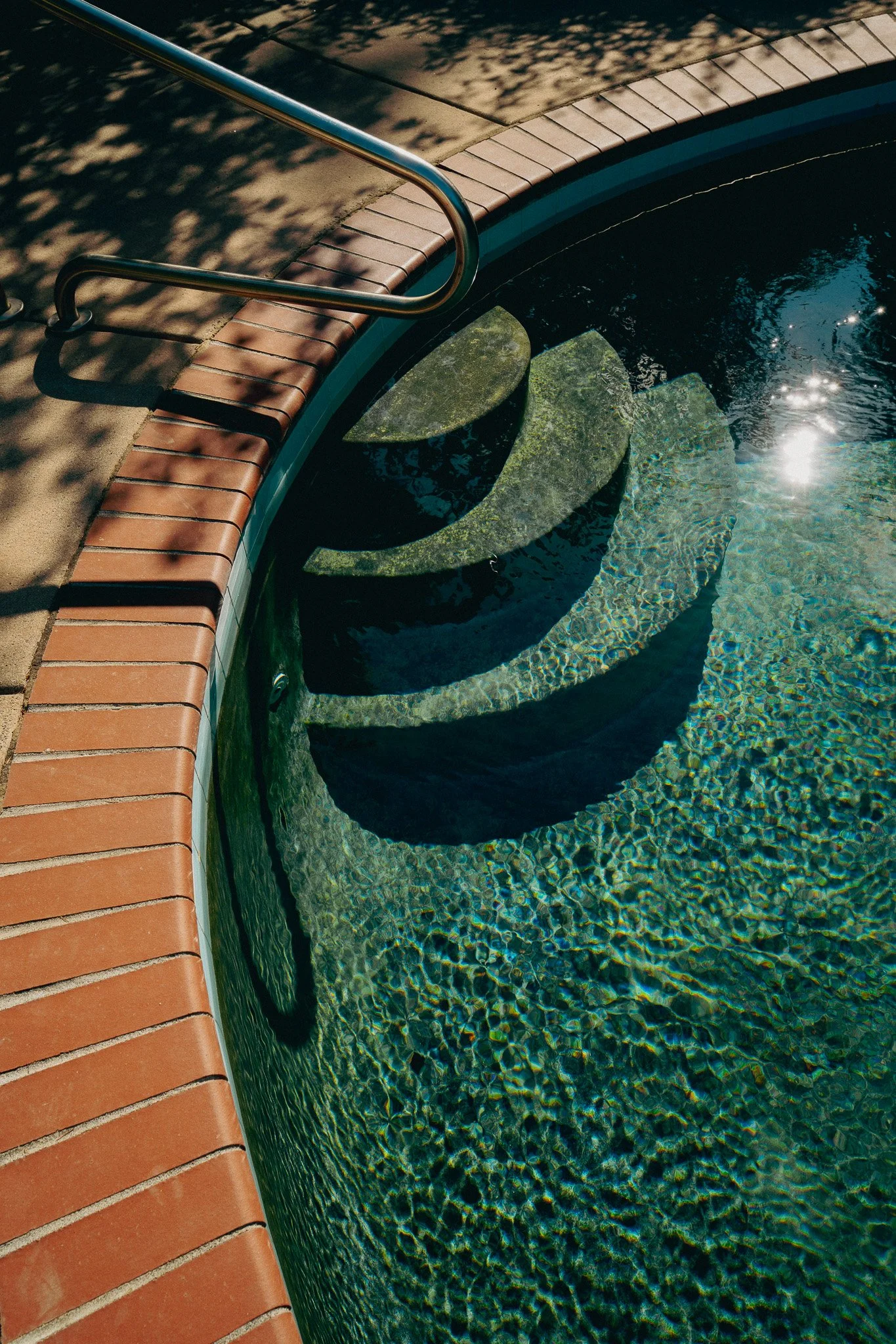 Close-up of a swimming pool's edge with a metal handrail, brick coping, and sunlight reflecting off the water, creating shadows and sparkles.