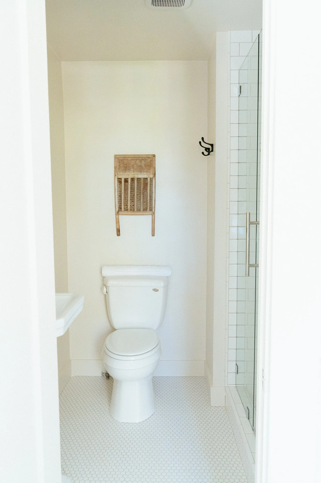 A small bathroom with white walls, a white toilet, a wooden shelf mounted on the wall above the toilet, a shower with a glass door, and a black towel hook on the wall.