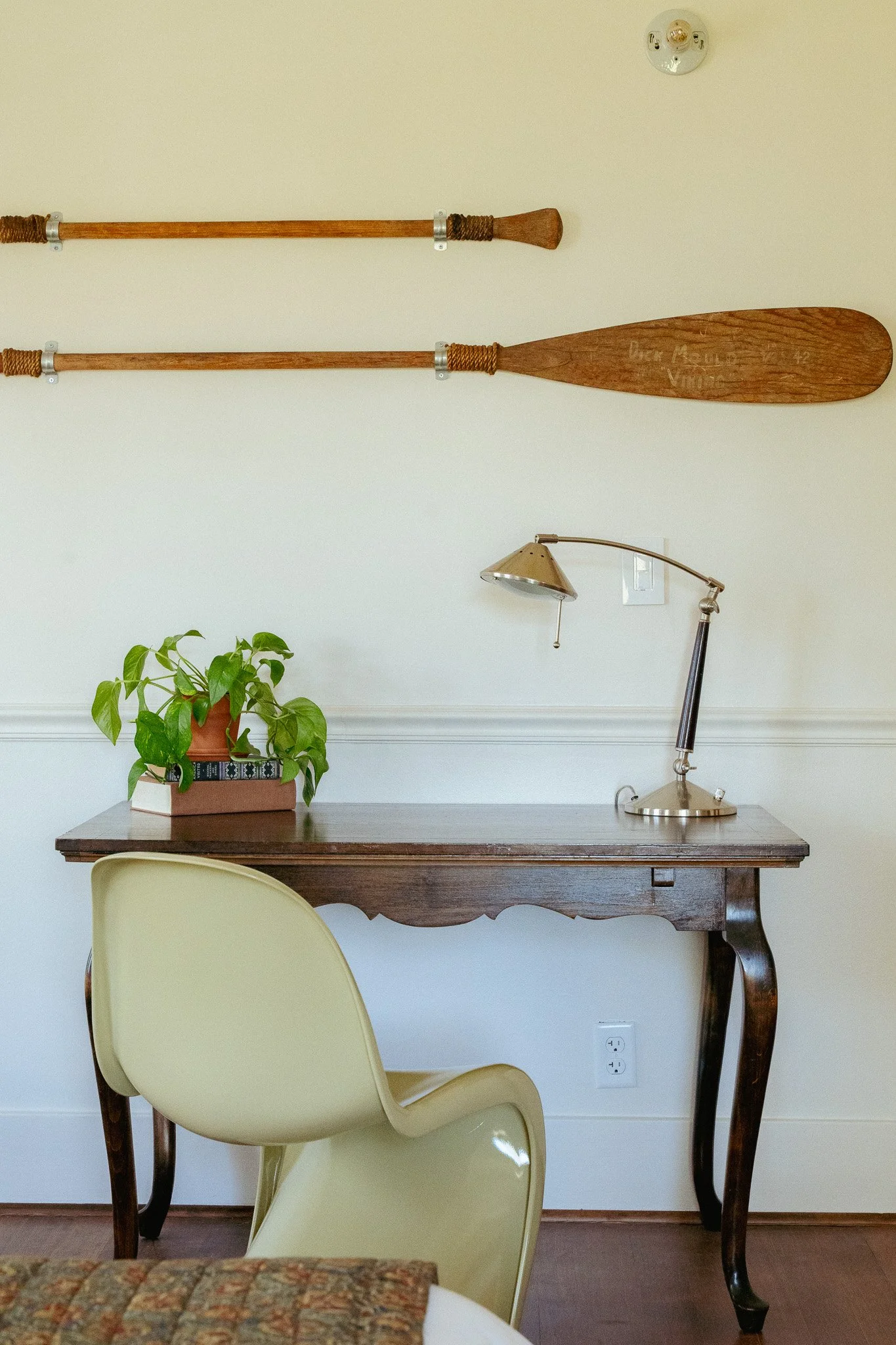 A wall with two wooden paddles hanging horizontally, with the larger paddle on the bottom and the smaller paddle on the top. Below the paddles, there is a wooden table with a potted plant and some books, and a yellow chair in front of the table. To t