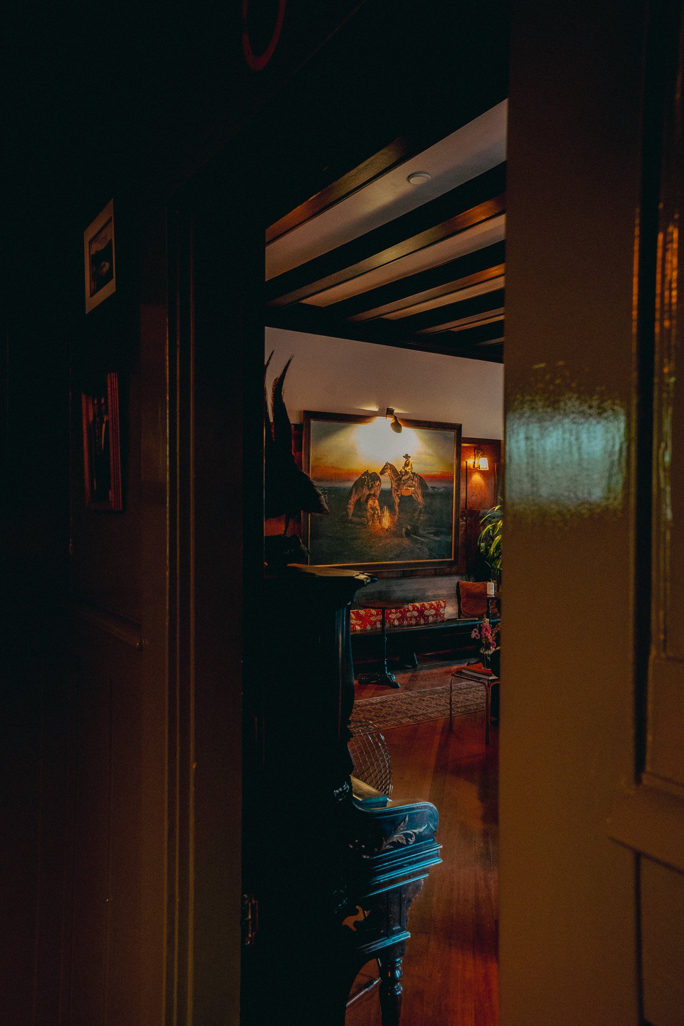 A dimly lit room with a painting of horses at sunset on the back wall, a small light fixture, a bench with a red and gold pillow, and a decorative rooster on a black piece of furniture.