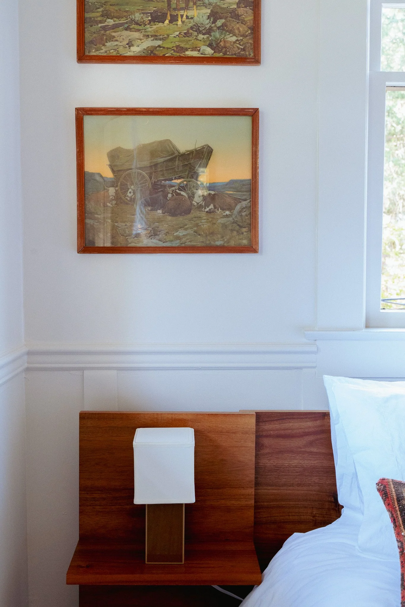 A bedroom nightstand with a white lamp, a bed with white sheets and pillows, a window with greenery outside, and framed artwork on the wall.