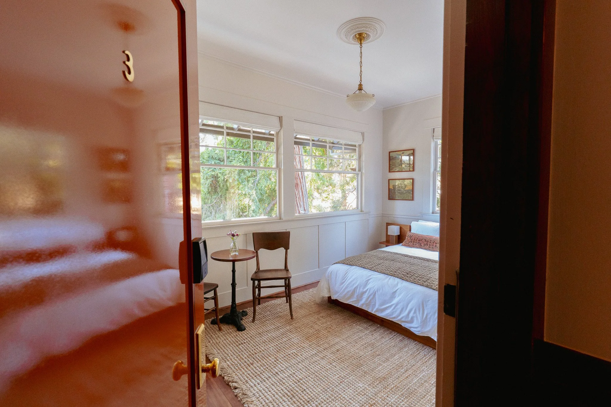 View of a cozy bedroom with a large window, a bed with a patterned bedspread, a small round table with a flower vase, two chairs, and framed pictures on the wall, seen through a slightly open door.