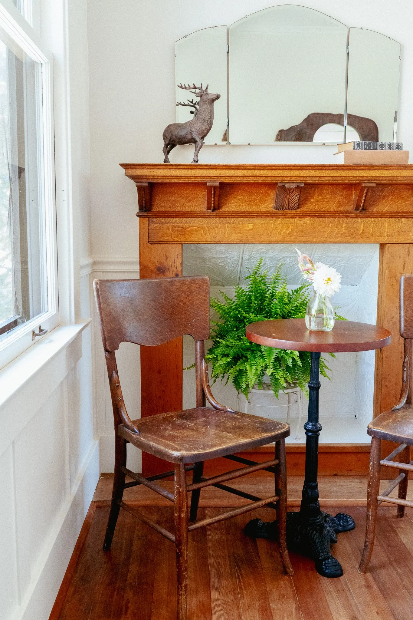 Interior of a cozy room with a wooden mantle, a vintage wooden chair, a small black table with a glass vase of white flowers, green ferns, and a large mirror on the wall reflecting part of the room.