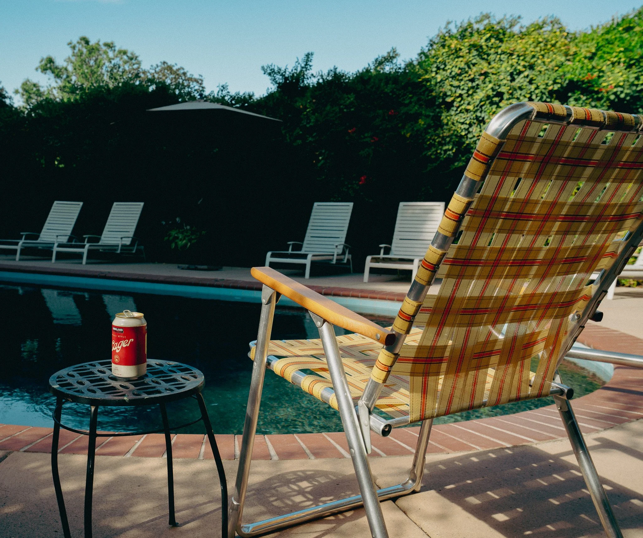 Poolside scene with a plaid patio chair in the foreground, a small round table with a can of soda, and a swimming pool with lounge chairs in the background, surrounded by lush green trees under a clear blue sky.