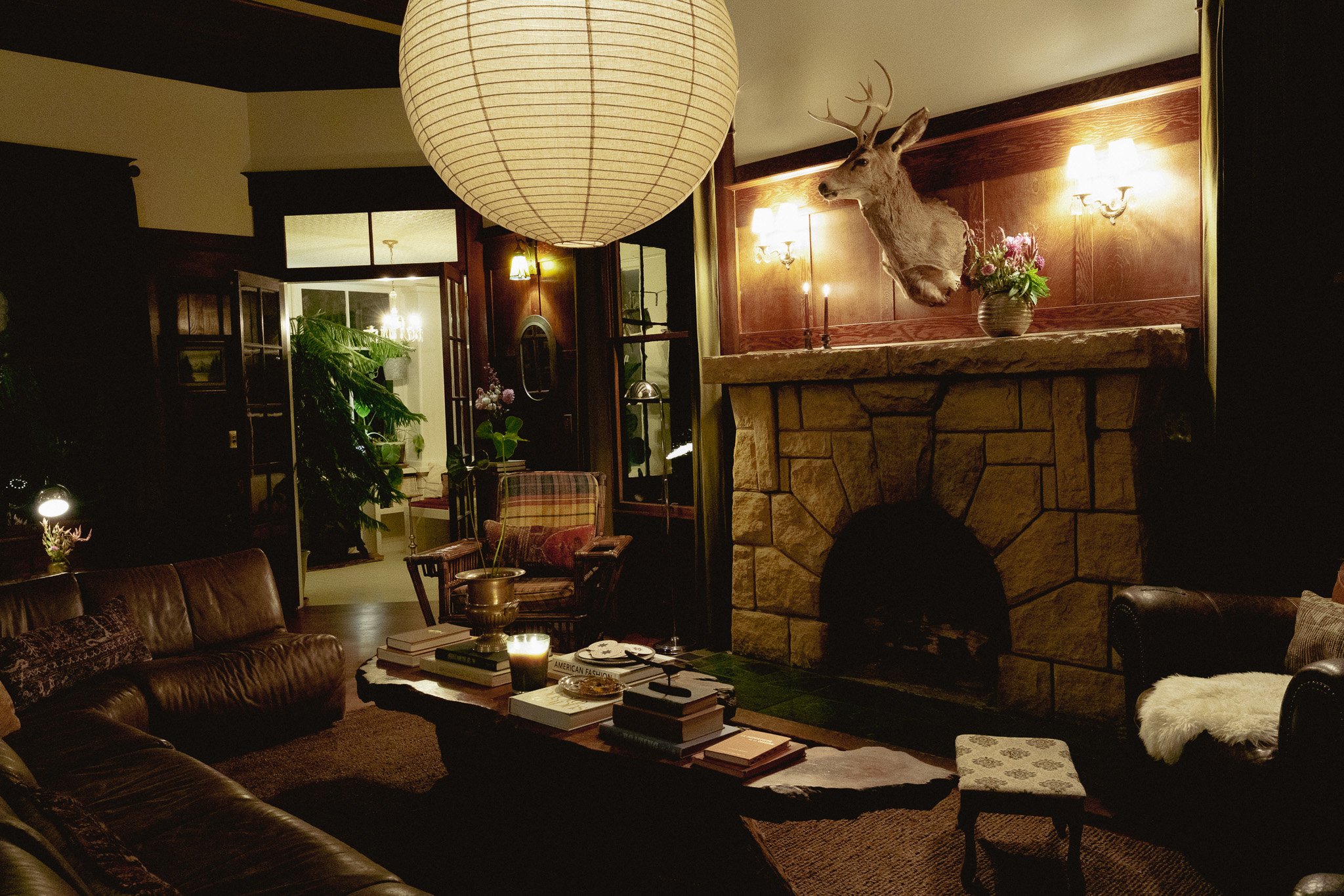 A cozy living room with a stone fireplace topped by a mounted deer head and a vase of flowers. The room is warmly lit with wall sconces and features a paper lantern hanging from the ceiling, a leather sofa, a stack of books on a coffee table, and large green plants.